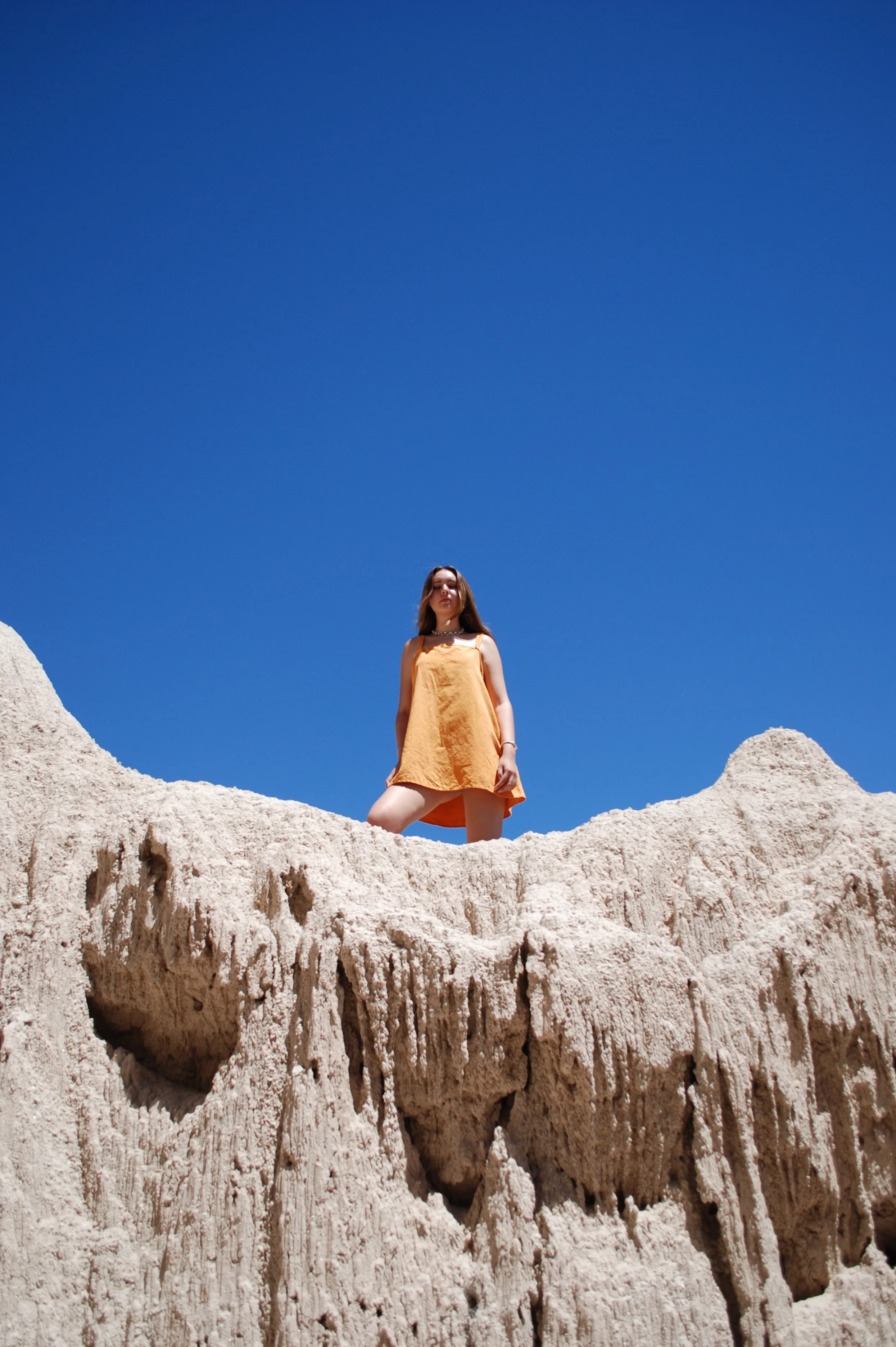 A woman in a yellow dress standing on a textured white rock formation against a clear blue sky.