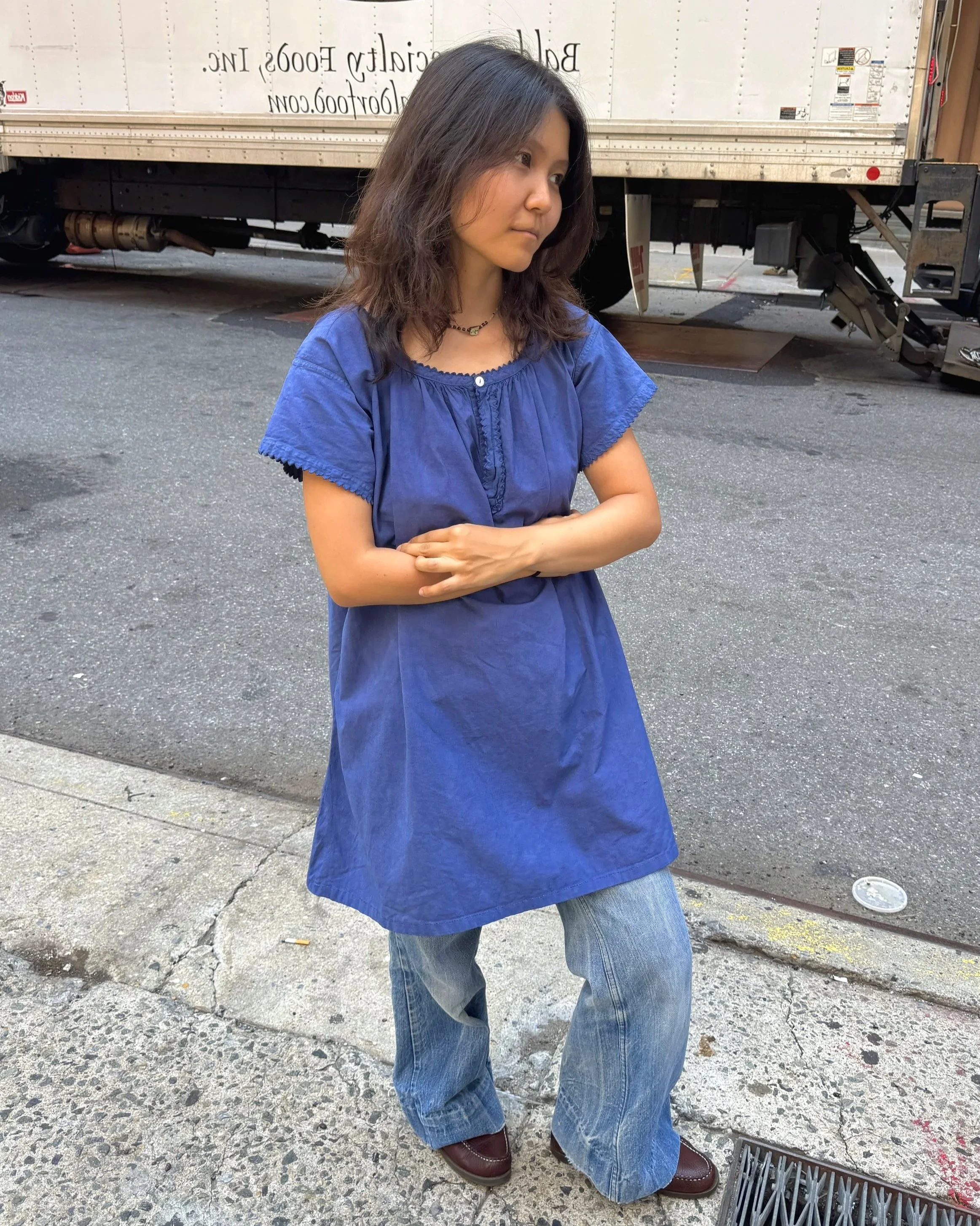 A woman with shoulder-length dark hair wearing a loose-fitting blue dress over jeans, standing outdoors on a concrete sidewalk near a large truck. She has her arms crossed and is wearing brown boots. The background includes the side of the truck and street elements.