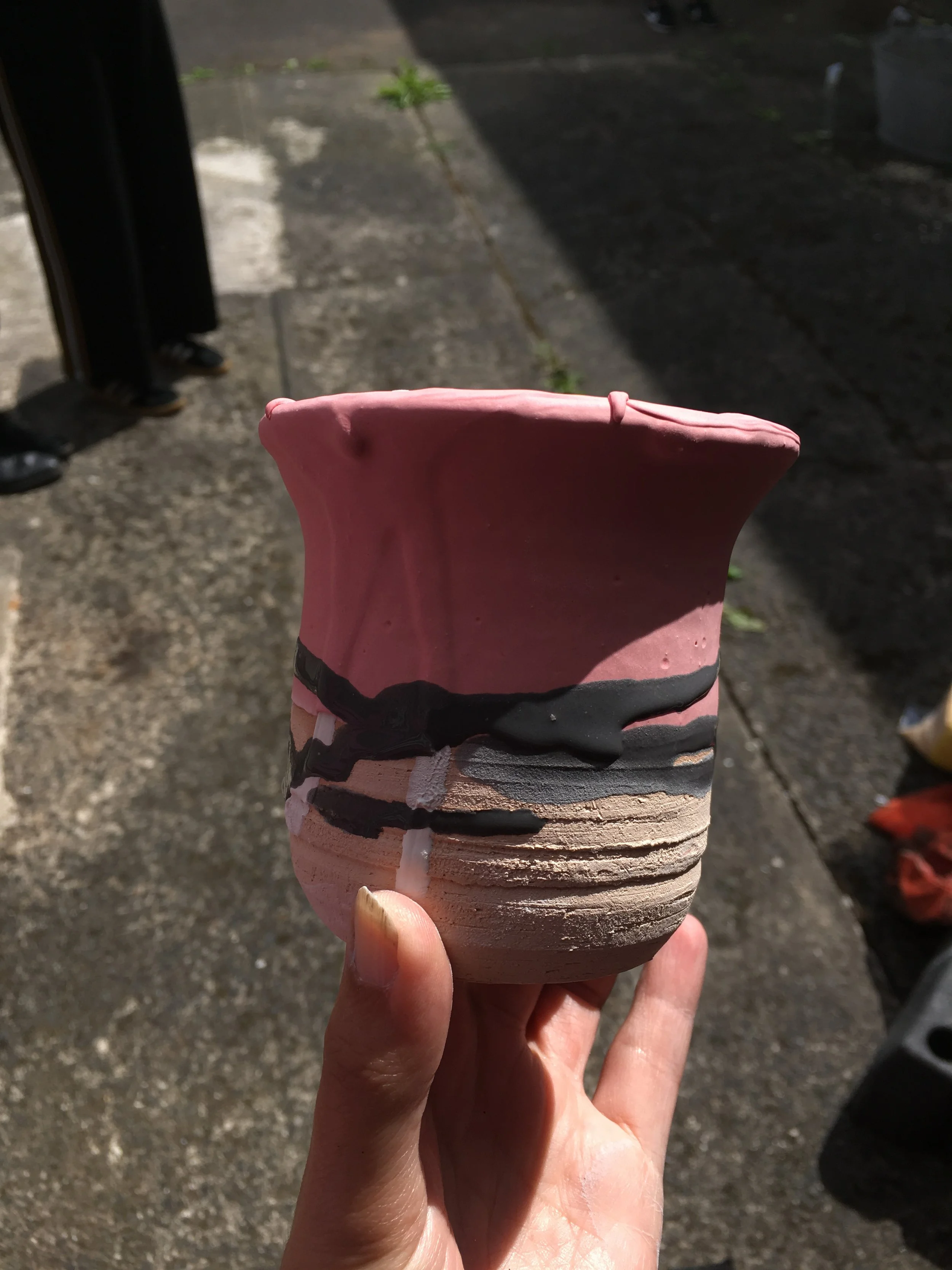 Close-up of a hand holding a pink and beige ice cream cone with chocolate drizzle, outdoors on pavement, with people in the background.