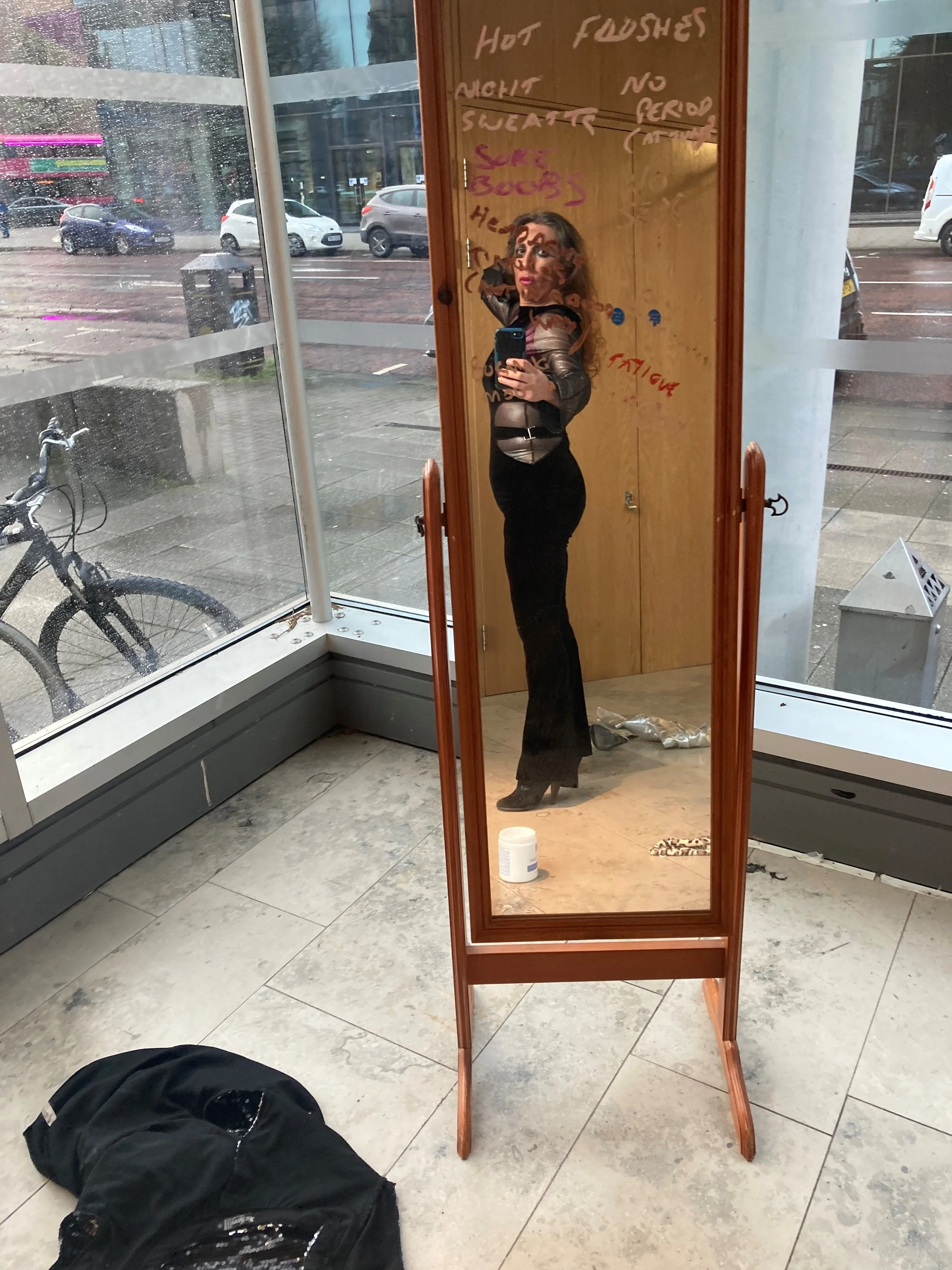 A woman taking a selfie in a tall, wooden-framed mirror inside a store, with rain outside visible through large glass windows. There is a bicycle, a backpack, and some items on the floor inside the store.