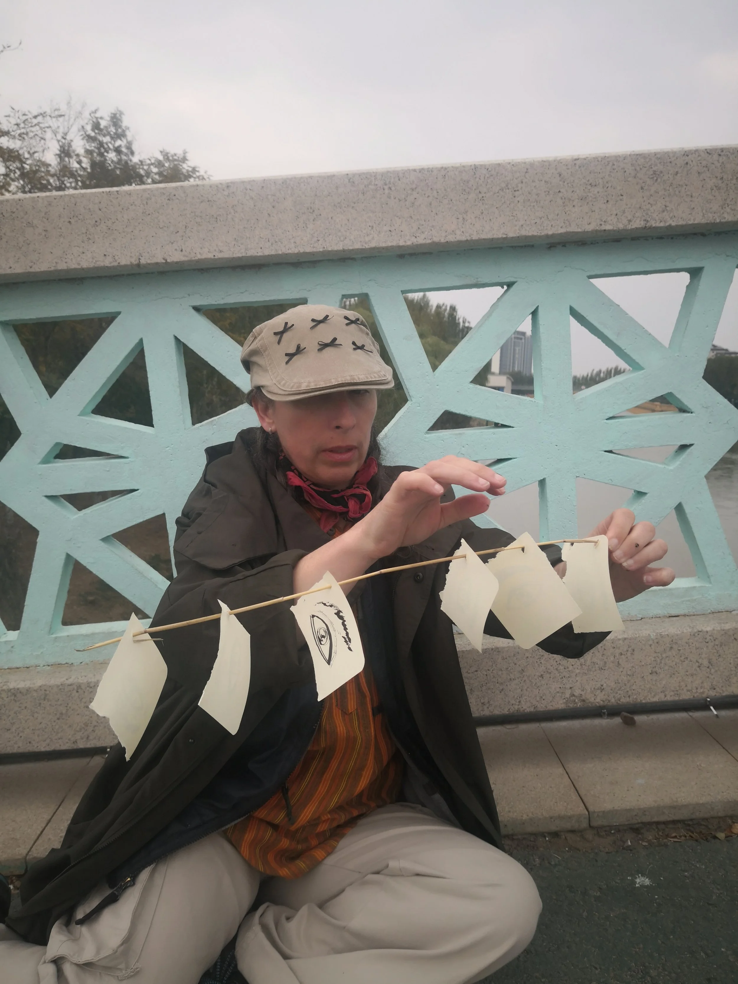 A person sitting on a bench, wearing a beige hat with black X's, a dark jacket, and khaki pants, holding a string of white paper flags with a drawing of an eye, with a light blue decorative fence in the background.