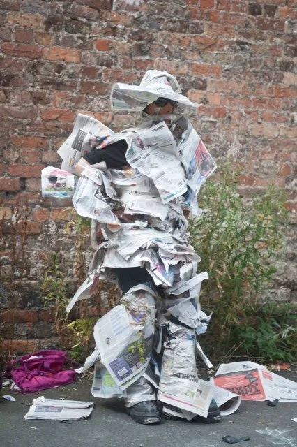 Person dressed in a suit made of crumpled newspapers standing against a brick wall.