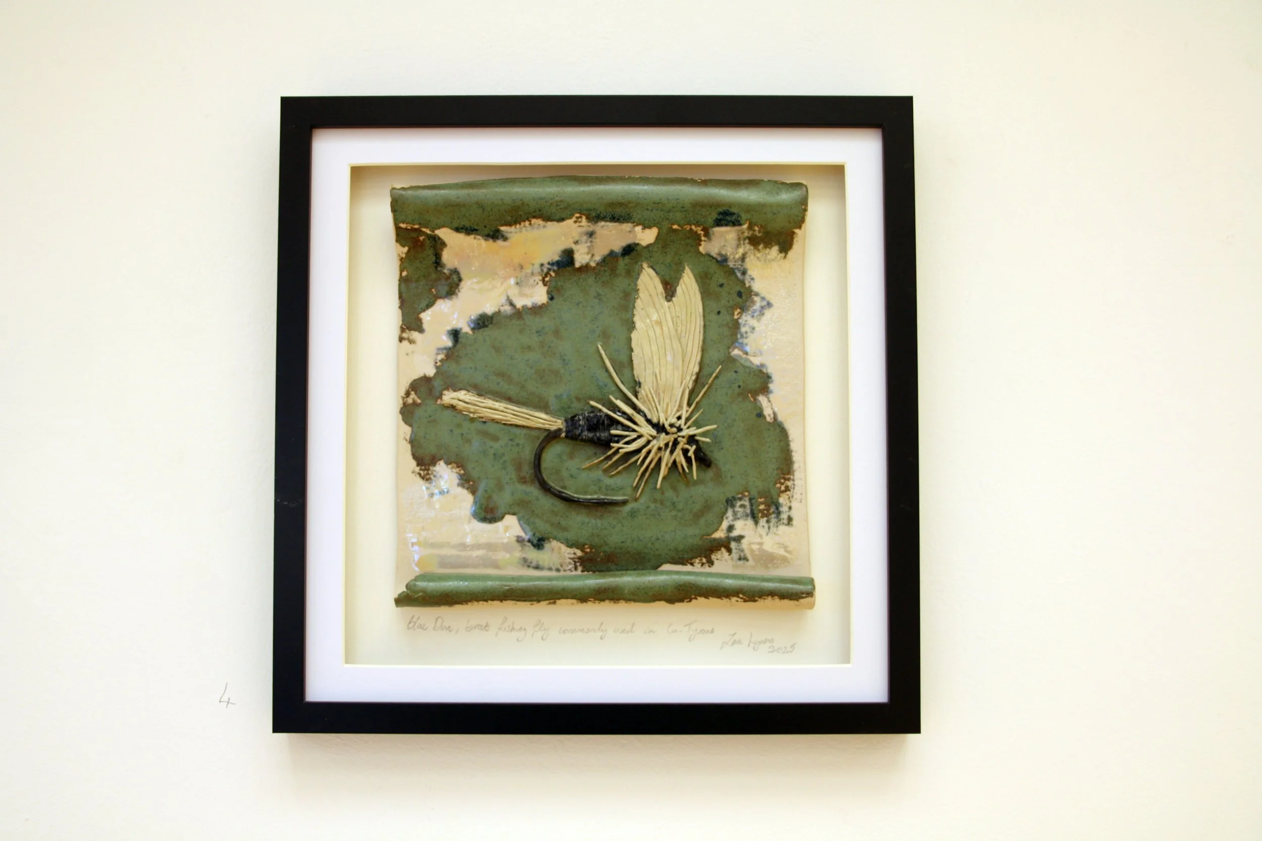 Framed artwork depicting a fishing lure with a butterfly wing design on a greenish background hanging on a white wall.