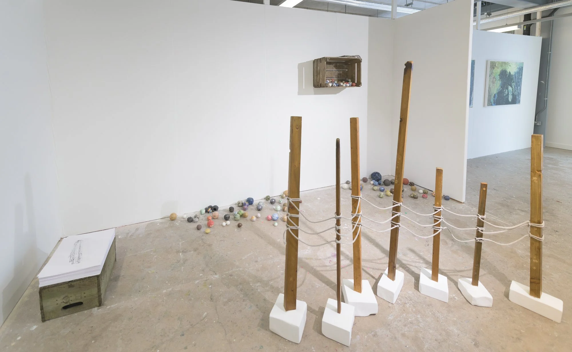 An art installation in a gallery featuring a wooden barrier with white bases and vertical wooden poles, along with scattered colorful balls on the floor. A white wall with a wooden box containing small objects and two paintings are visible in the bac