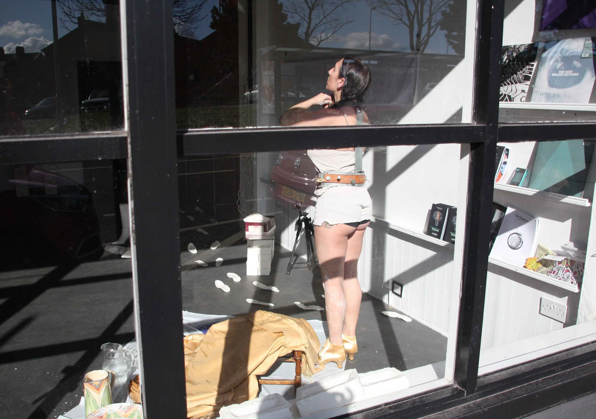 A woman with dark hair, wearing a black top and white shorts, is standing inside a room with a window, adjusting her hair. The room has various books and items on shelves and tables, with sunlight streaming in through the windows. Outside, parked car
