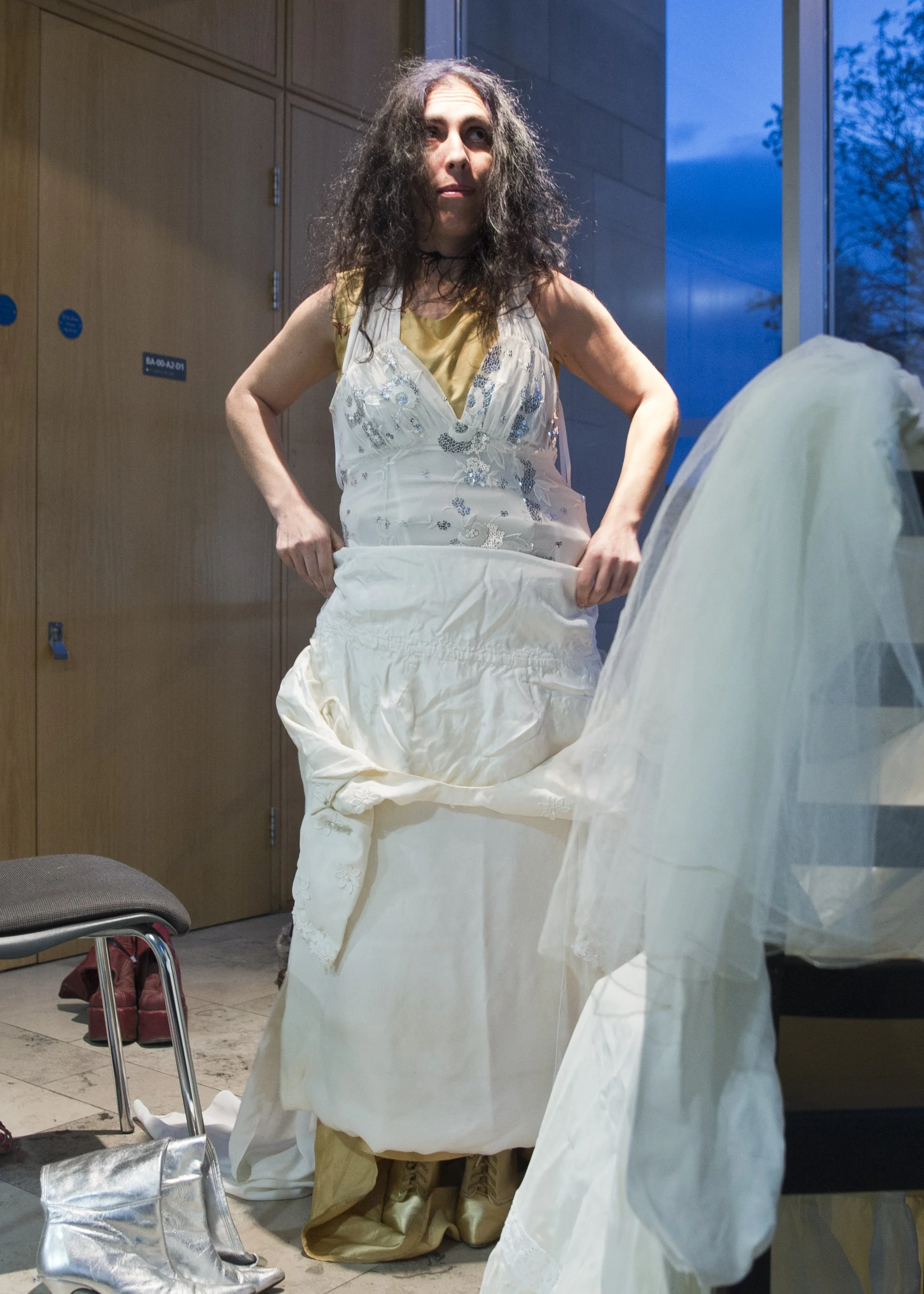A person with long, curly hair wearing a yellow shirt and a white dress with blue embroidery, holding a white fabric, standing indoors near large windows with trees outside, a chair with a bag and red shoes nearby.