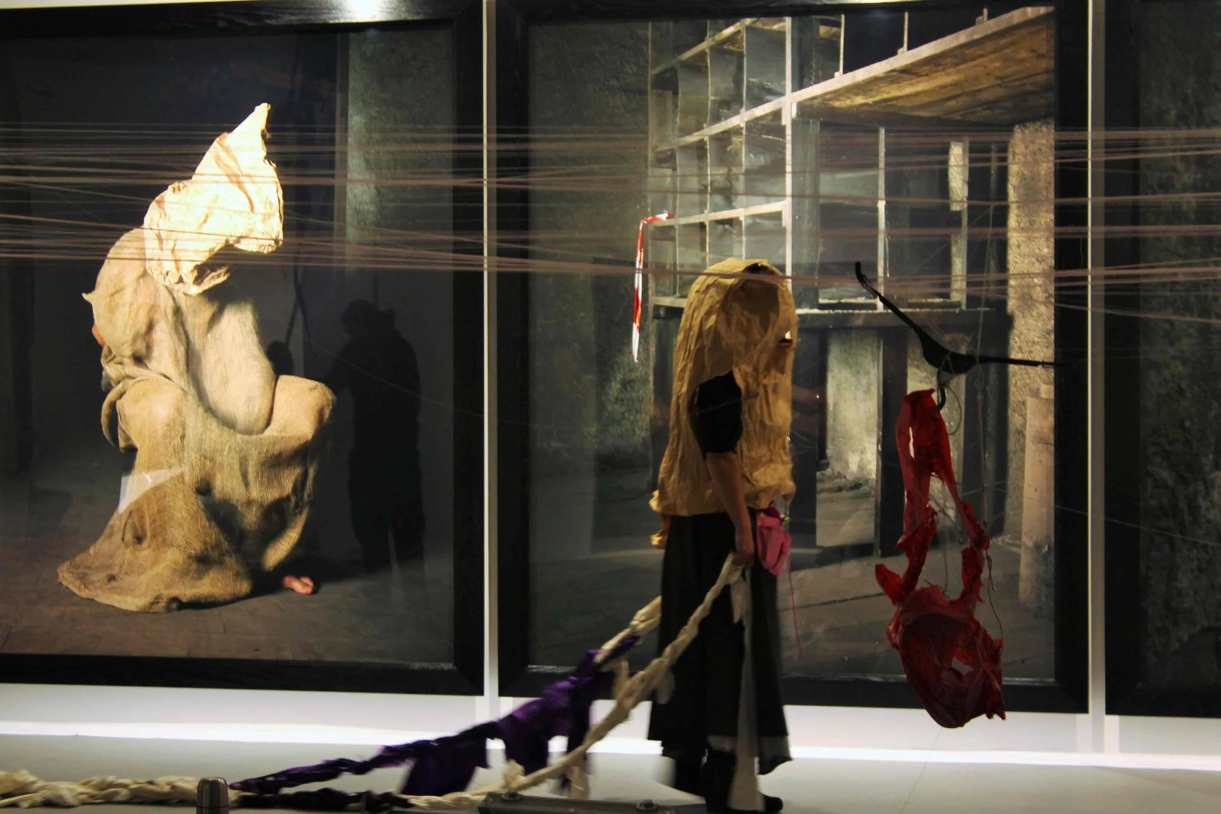 Art exhibit featuring large paper sculptures resembling a seated person and a head, with string and fabric elements, viewed through glass.