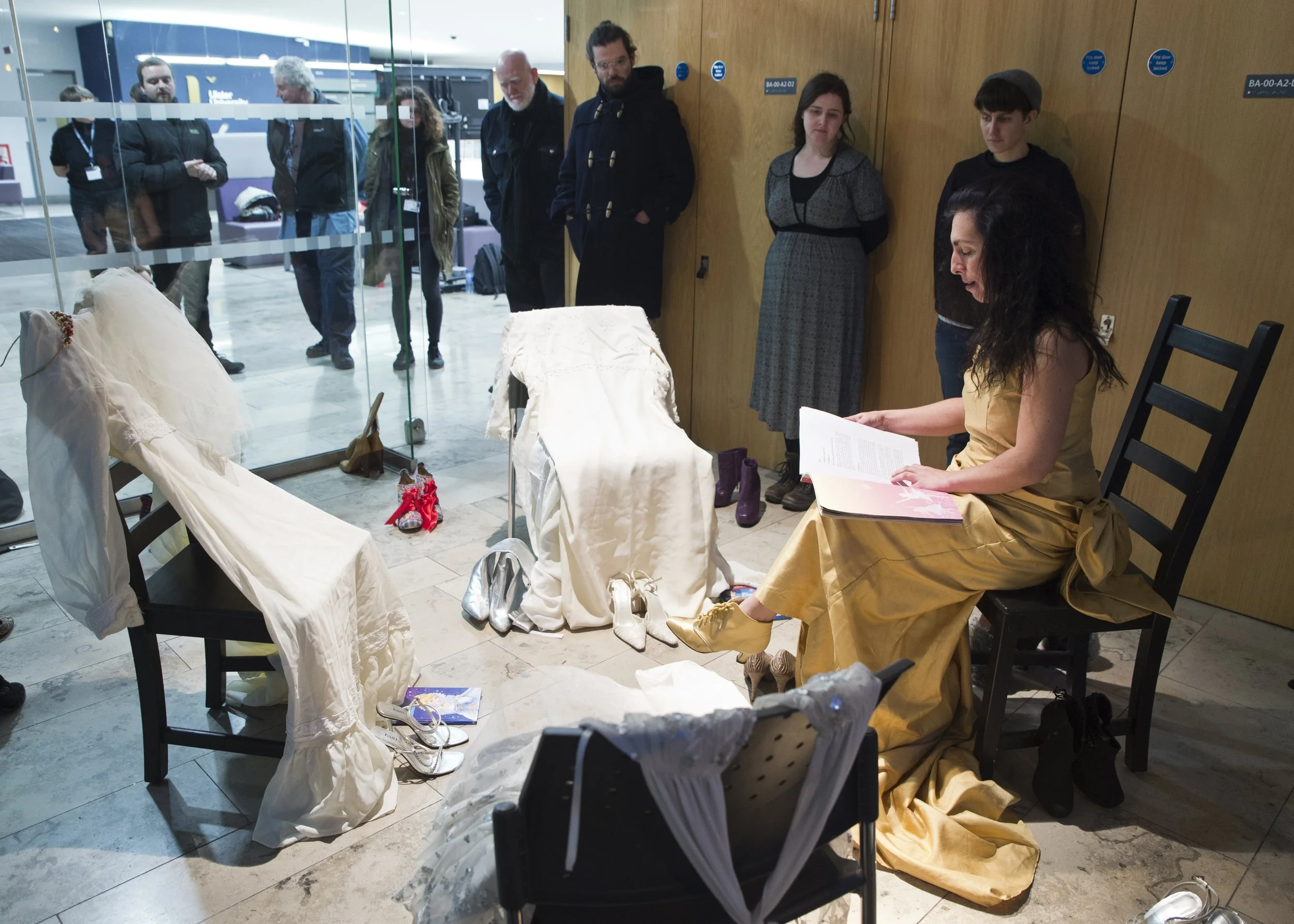 Group of people observing a display of wedding dresses and accessories in a room, with some seated and others standing behind a glass partition.