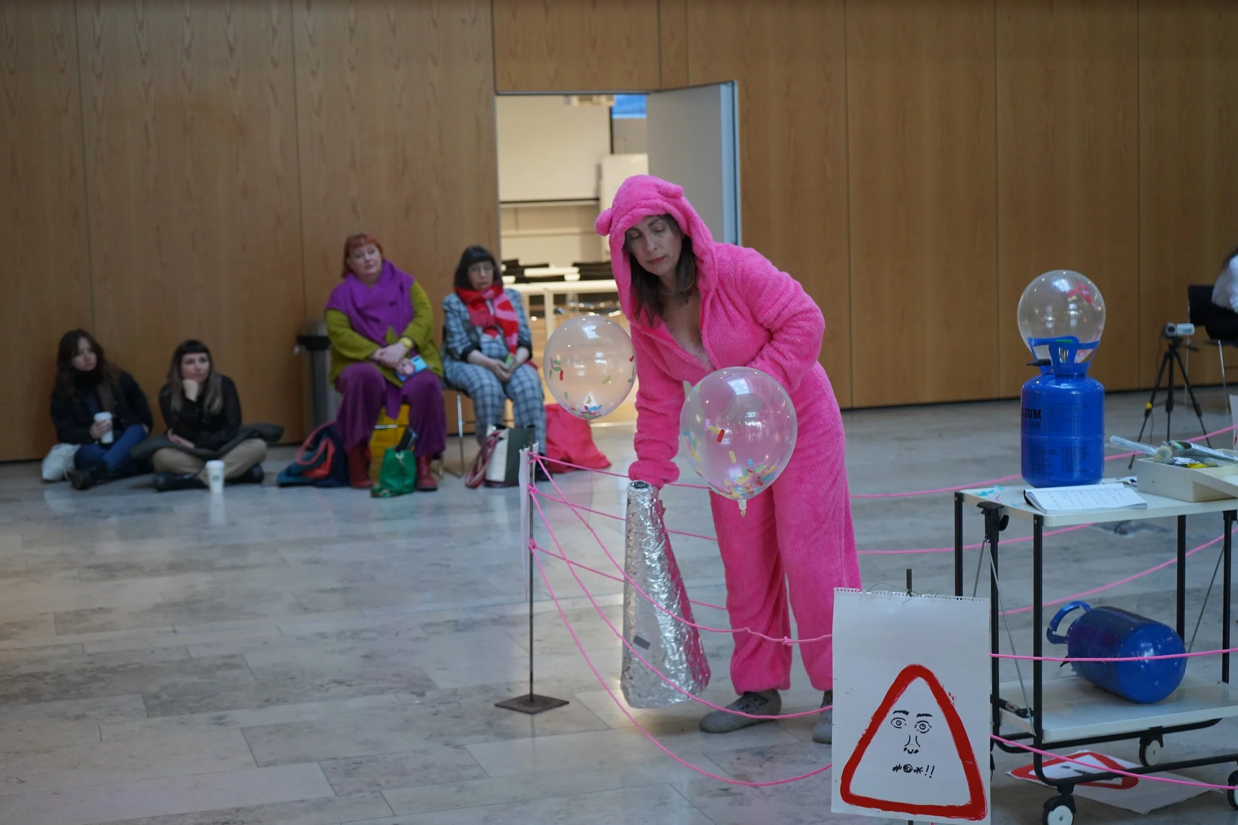 Woman in a pink fluffy costume with a hood that has ears, performing a science experiment with balloons and a cone in a public indoor space, while four women watch seated in the background.