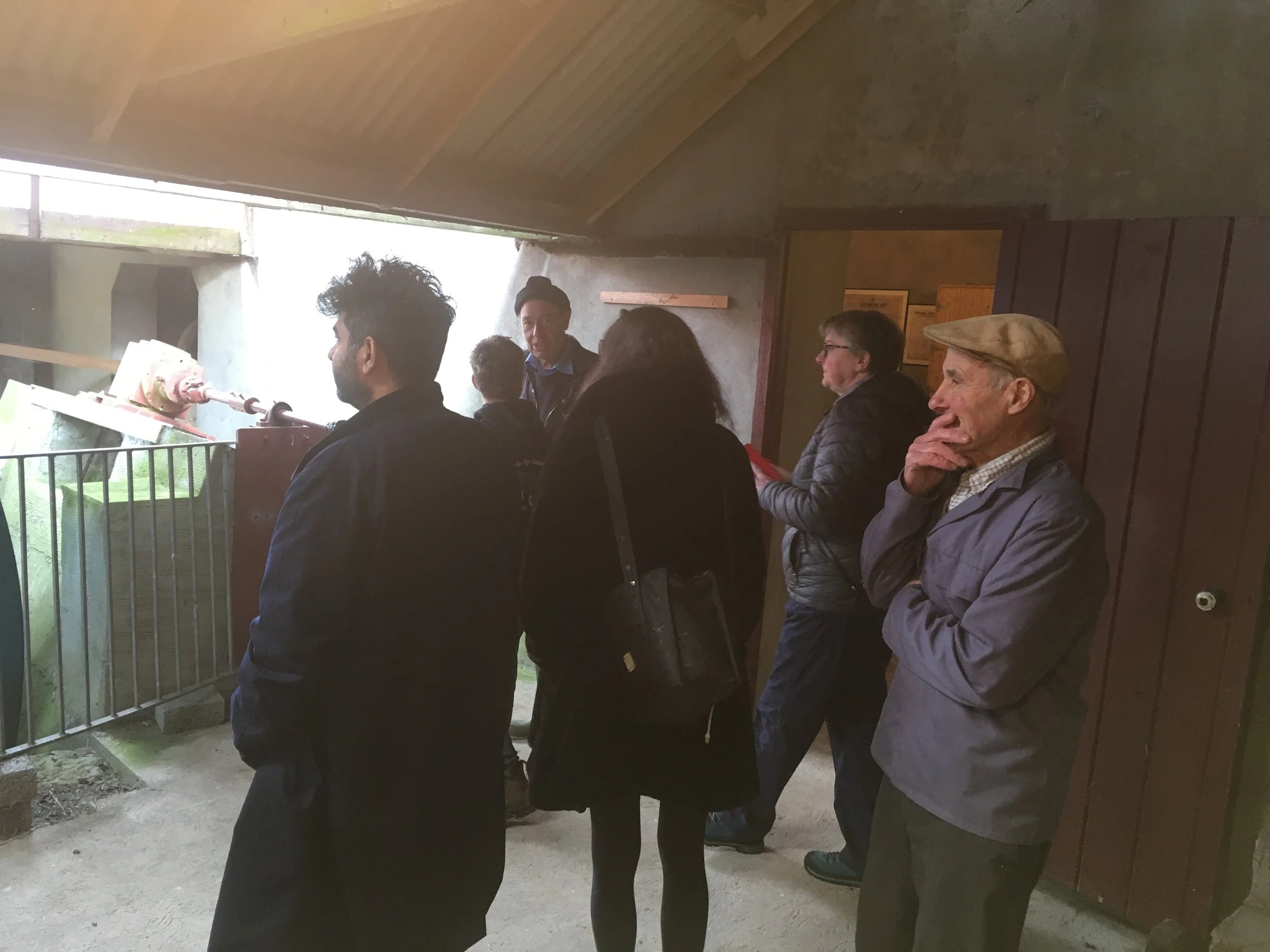 A group of people standing inside a building next to a railing, looking out through an opening to an outdoor area with grass. The people are engaged and appear to be listening or observing something.