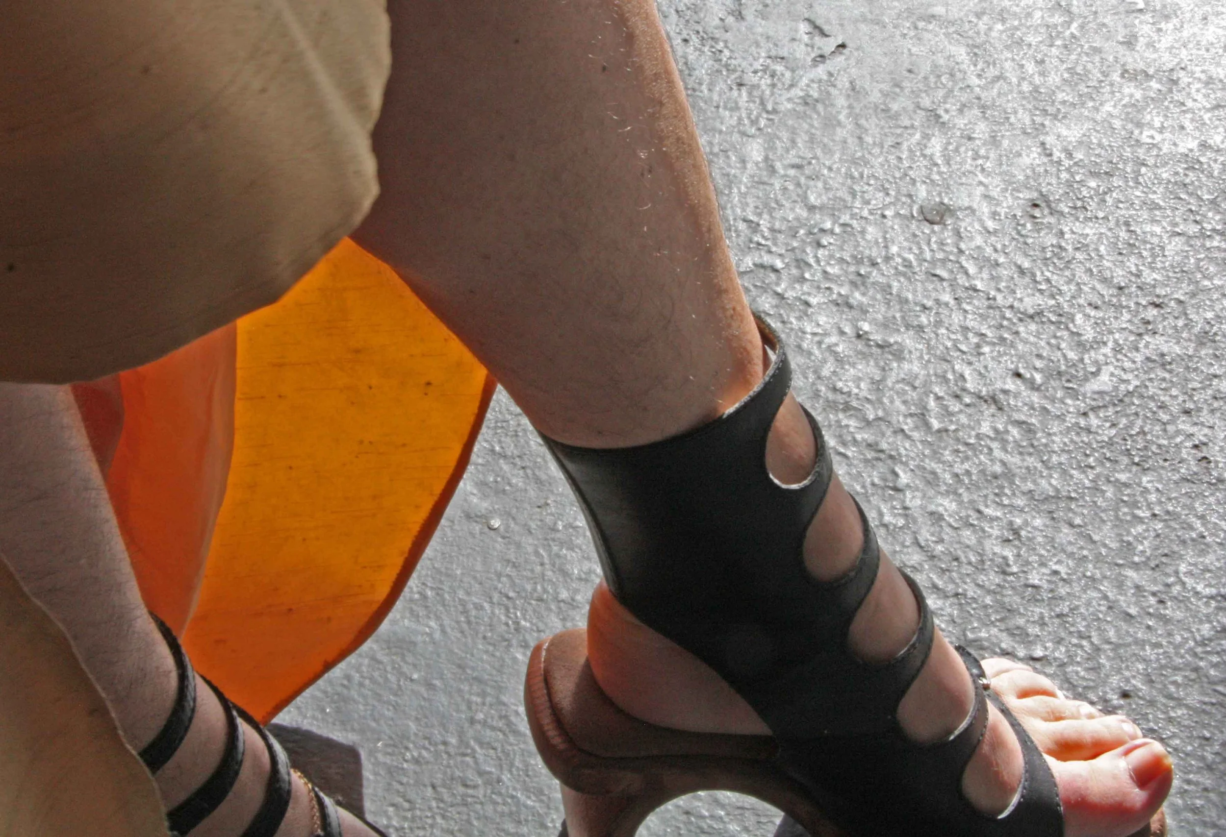 A person wearing black strappy sandals with rings on toes, sitting on an orange stool with legs crossed, on a concrete floor.
