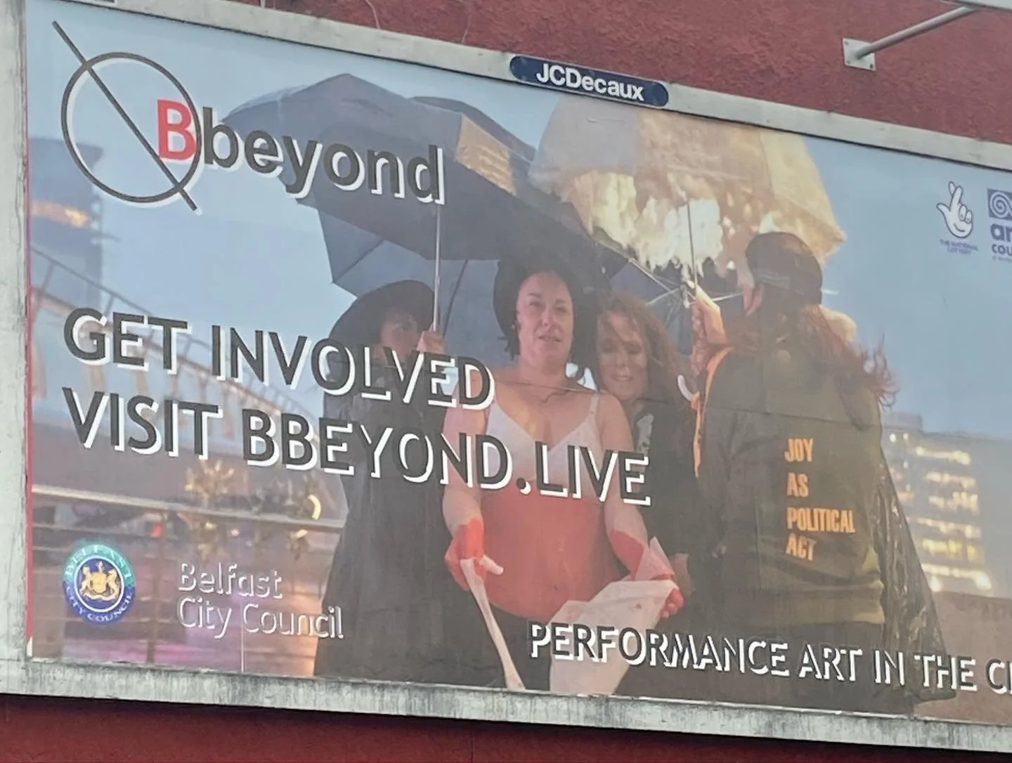 A banner with the logo of Belfast City Council, showing diverse women holding umbrellas, promoting community involvement and performance art, with text encouraging people to get involved and visit beyond.live.