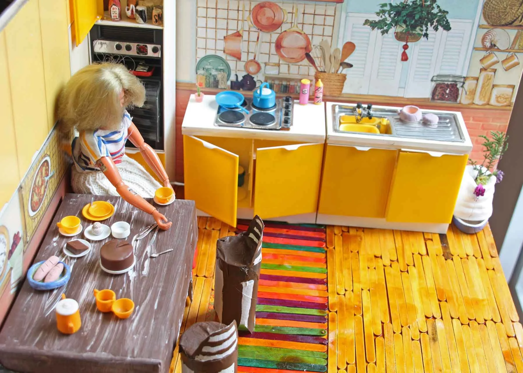 Miniature model of a colorful kitchen with yellow cabinets, a stove, a sink, a small dining table with toy dishes, a striped rug, and a wall decorated with kitchen-themed wallpaper.