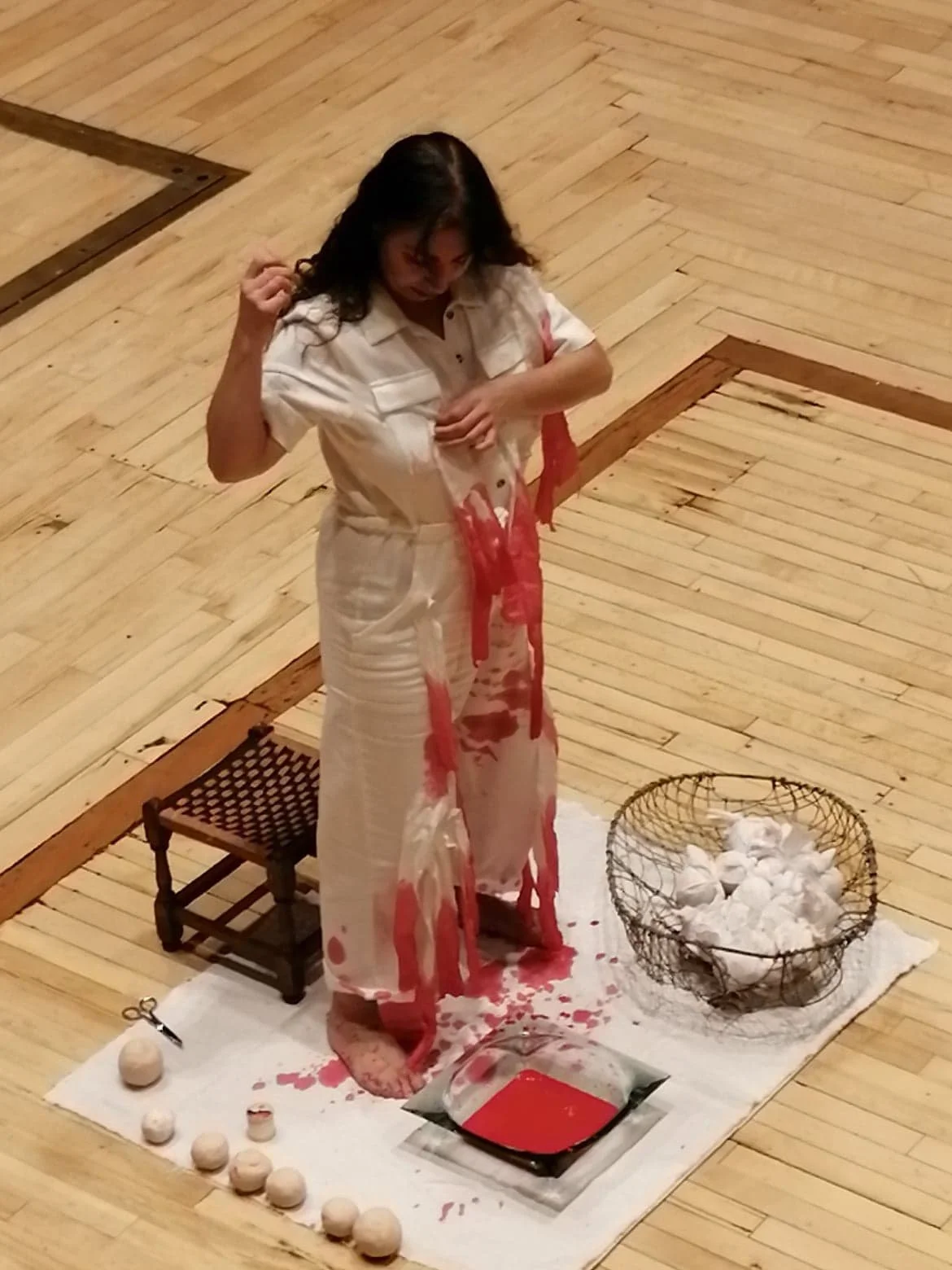 A woman in white clothes standing on a wooden floor with red paint on her clothes and body, holding her hair with her left hand. There is a small chair, a woven basket filled with white balls, and a white cloth beneath her. Blood-like red liquid is o