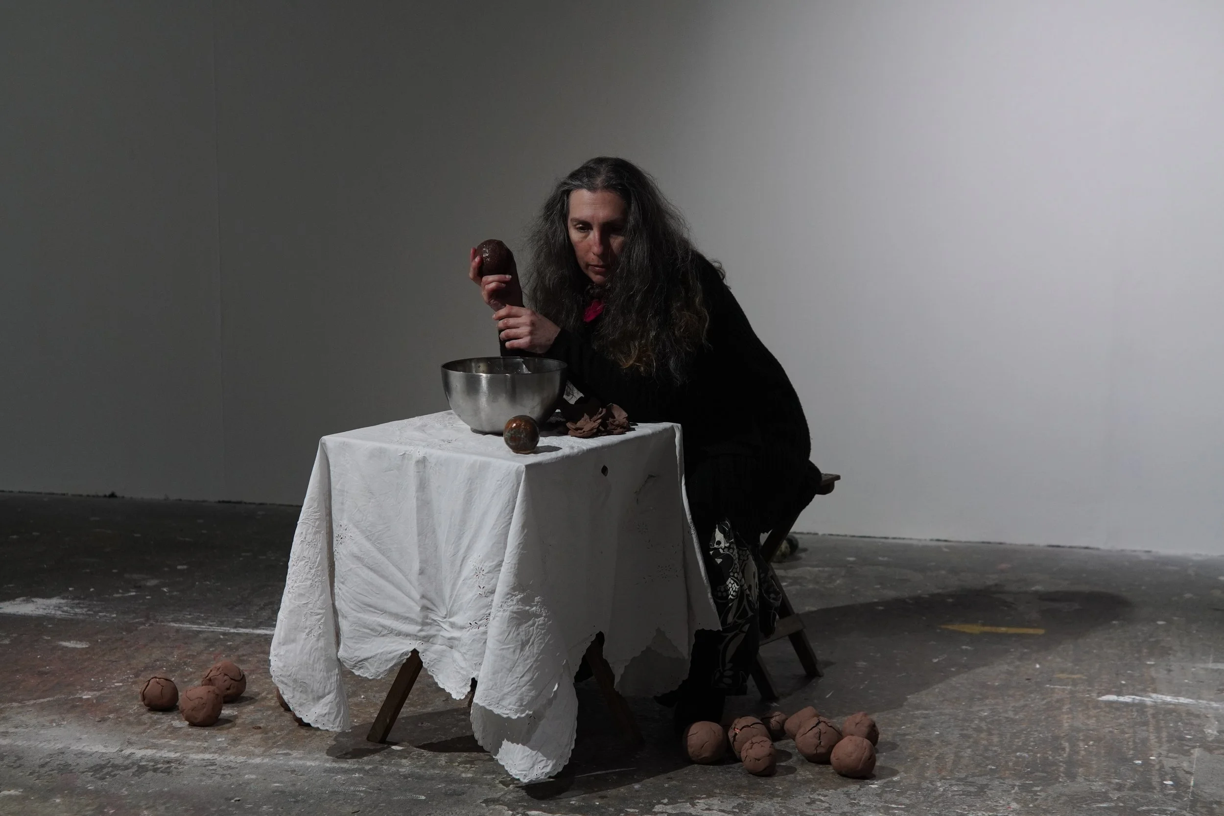 A woman with long dark hair wearing black, kneeling beside a table covered with a white tablecloth, holding a chocolate-colored egg in her right hand, with more eggs and a metal bowl on the table, and a few eggs scattered on the floor, against a plain white wall background.