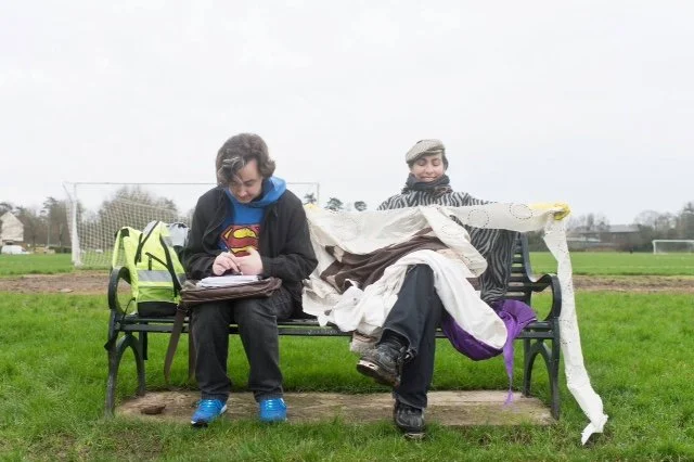 Two people sitting on a park bench, with one girl looking at a device and a boy covered in fabric or paper, wearing a newsboy cap, in a grassy field with soccer goals in the background.