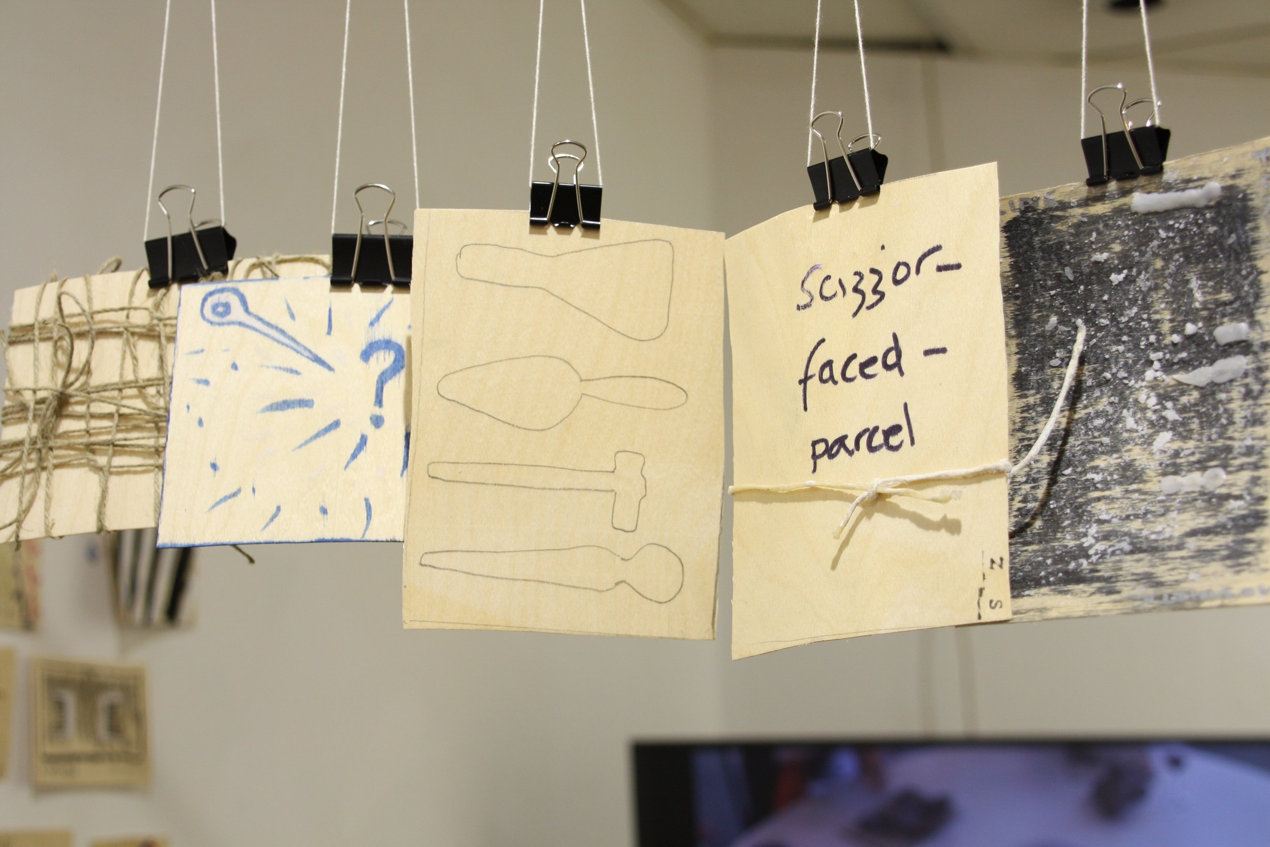 Several paper notes are clipped to a string with black binder clips, with sketches and handwriting on them. The notes include drawings of tools and handwritten notes, with one visible text reading 'scissors - faced - parcel'.