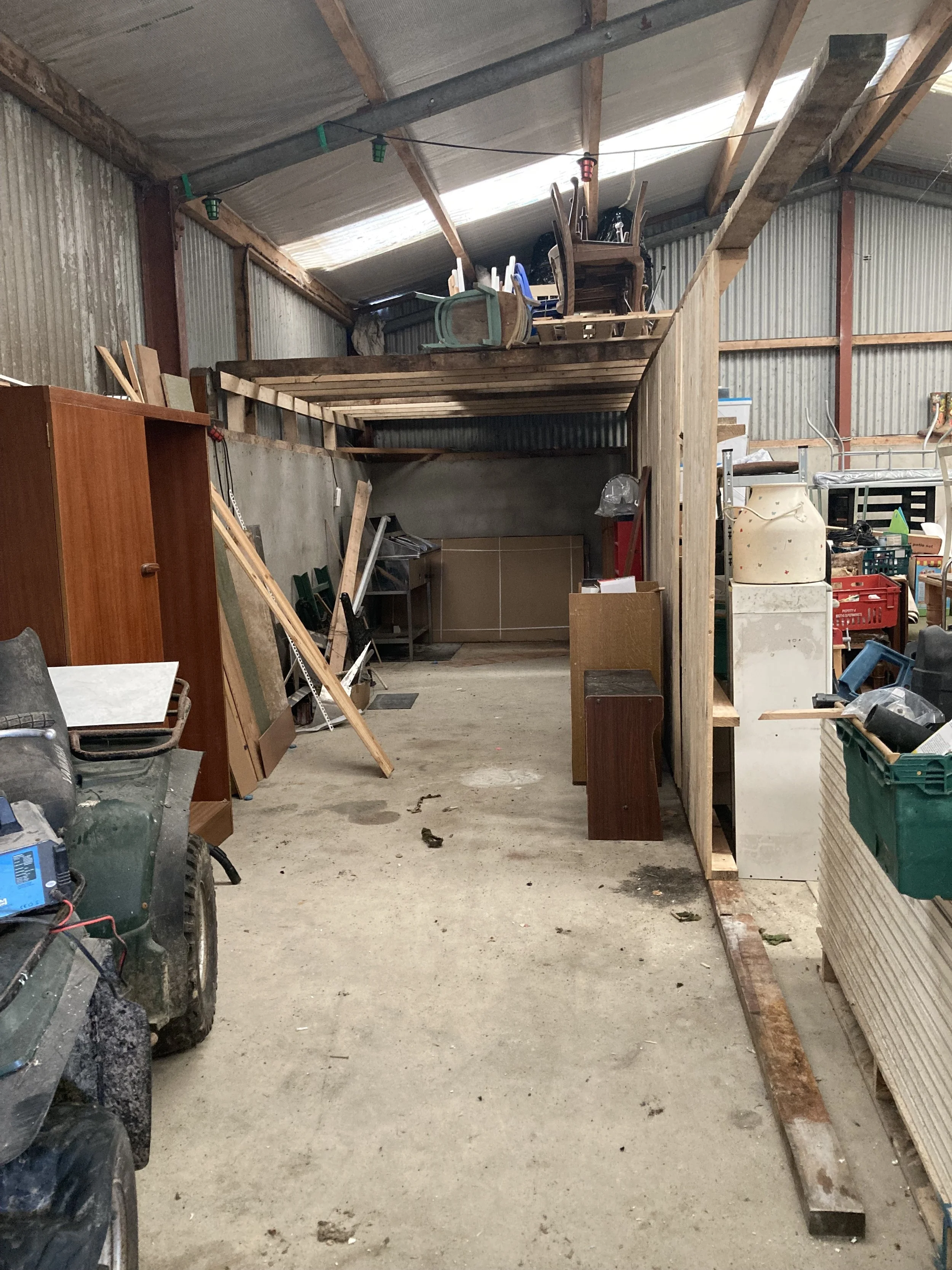 A cluttered garage with construction materials, furniture, and various tools. The upper level has chairs and a small table, while the lower level has a workbench, a cabinet, and a tractor tire. The walls are made of corrugated metal, and the ceiling has exposed wooden beams.