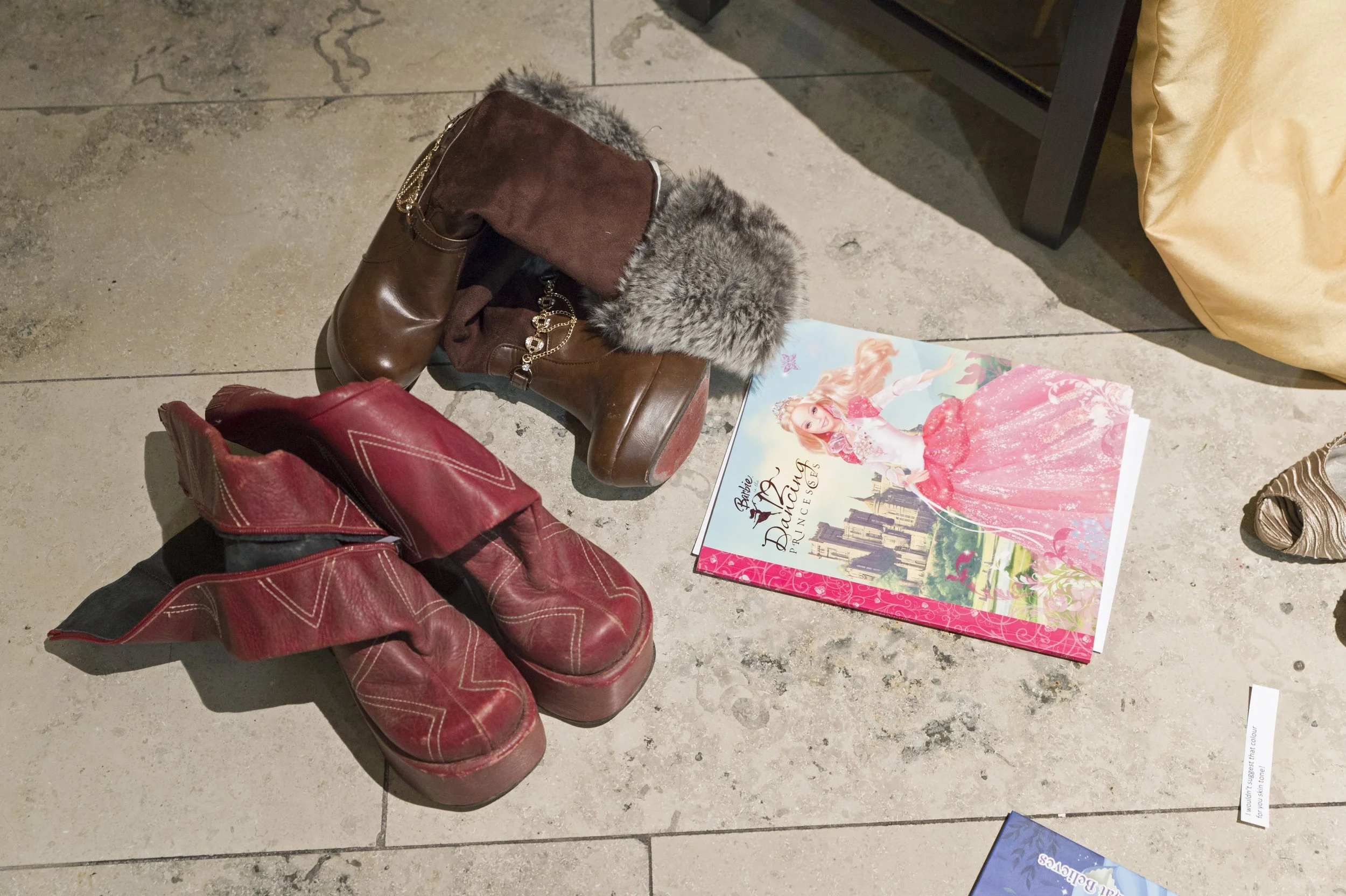 A pair of red and brown boots with faux fur lining and a chain detail, placed next to a red bag and a colorful book with a princess on the cover, on a tiled floor.