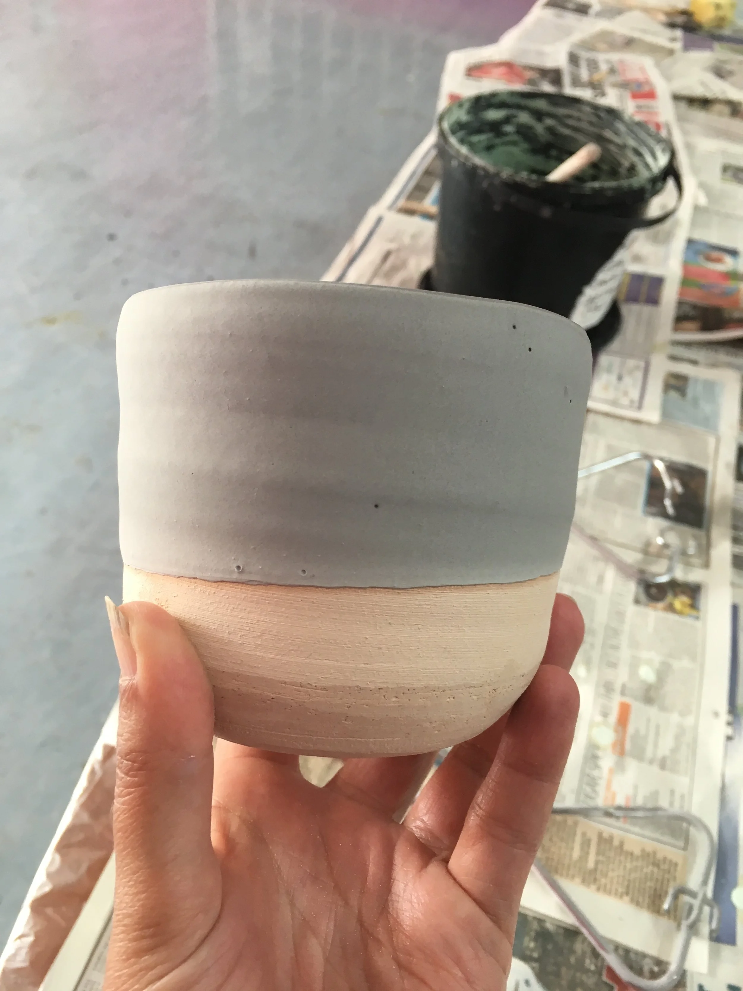 A person holding a blank, unpainted ceramic mug in the foreground, with a small paintbrush inside a dark container and newspapers spread out in the background, indicating a pottery or painting workshop.