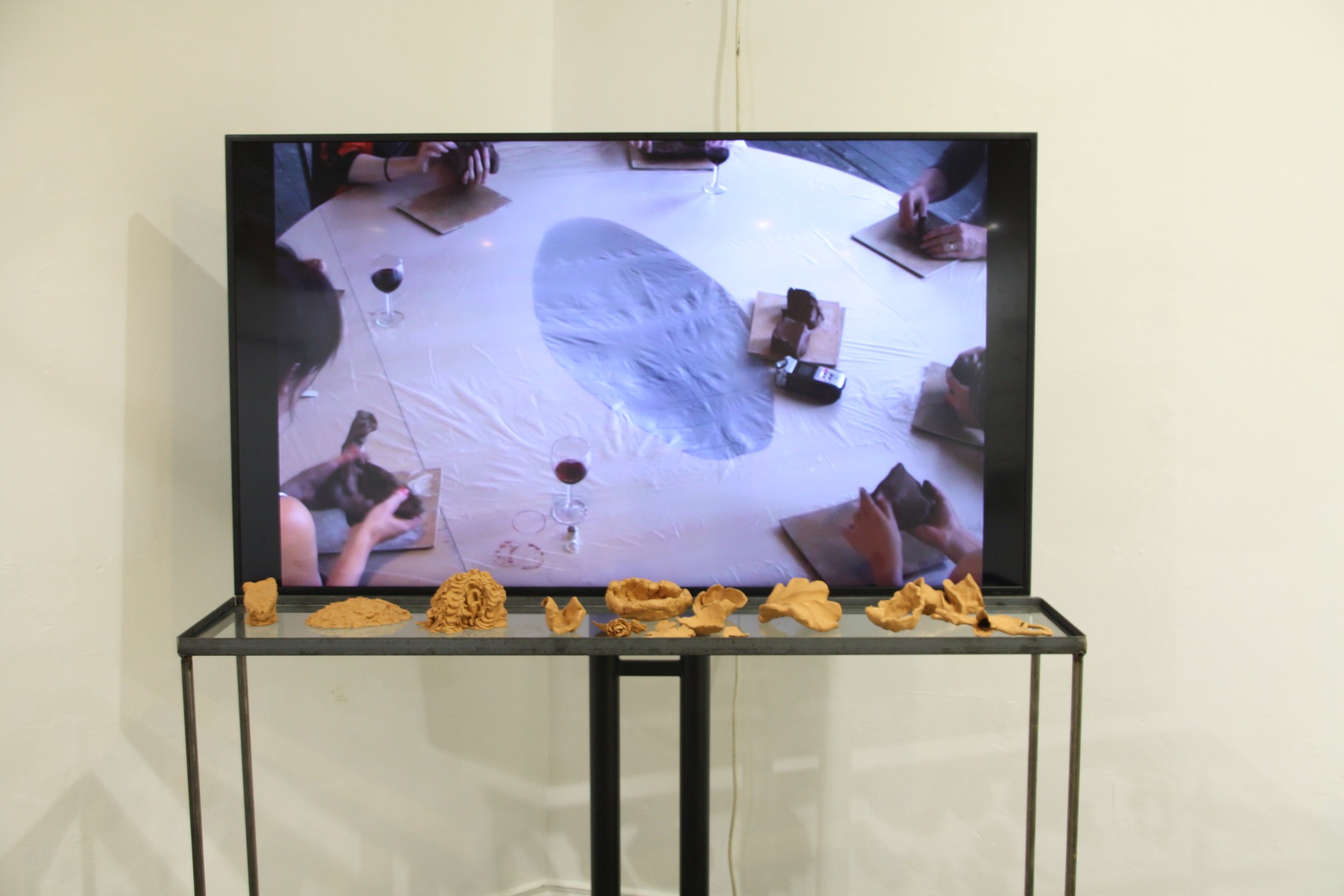 A television screen displaying a group of people with glasses of wine, sitting at a long table with papers and objects on top. The photo also shows an art display at the bottom with clay sculptures.