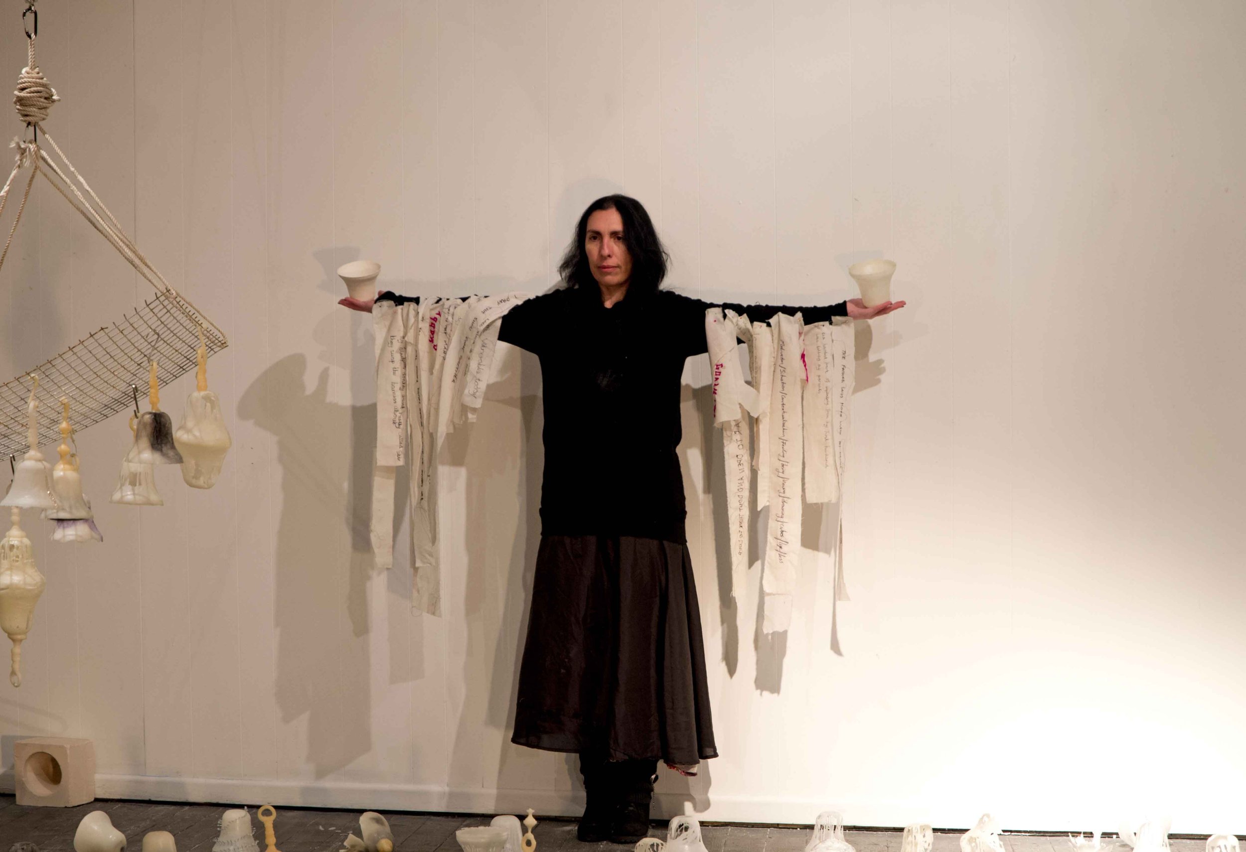 A woman in black clothing standing with arms extended, holding white ceramic objects on each hand, in front of a wall with hanging fabric strips with writings, surrounded by various ceramic pieces on the floor.