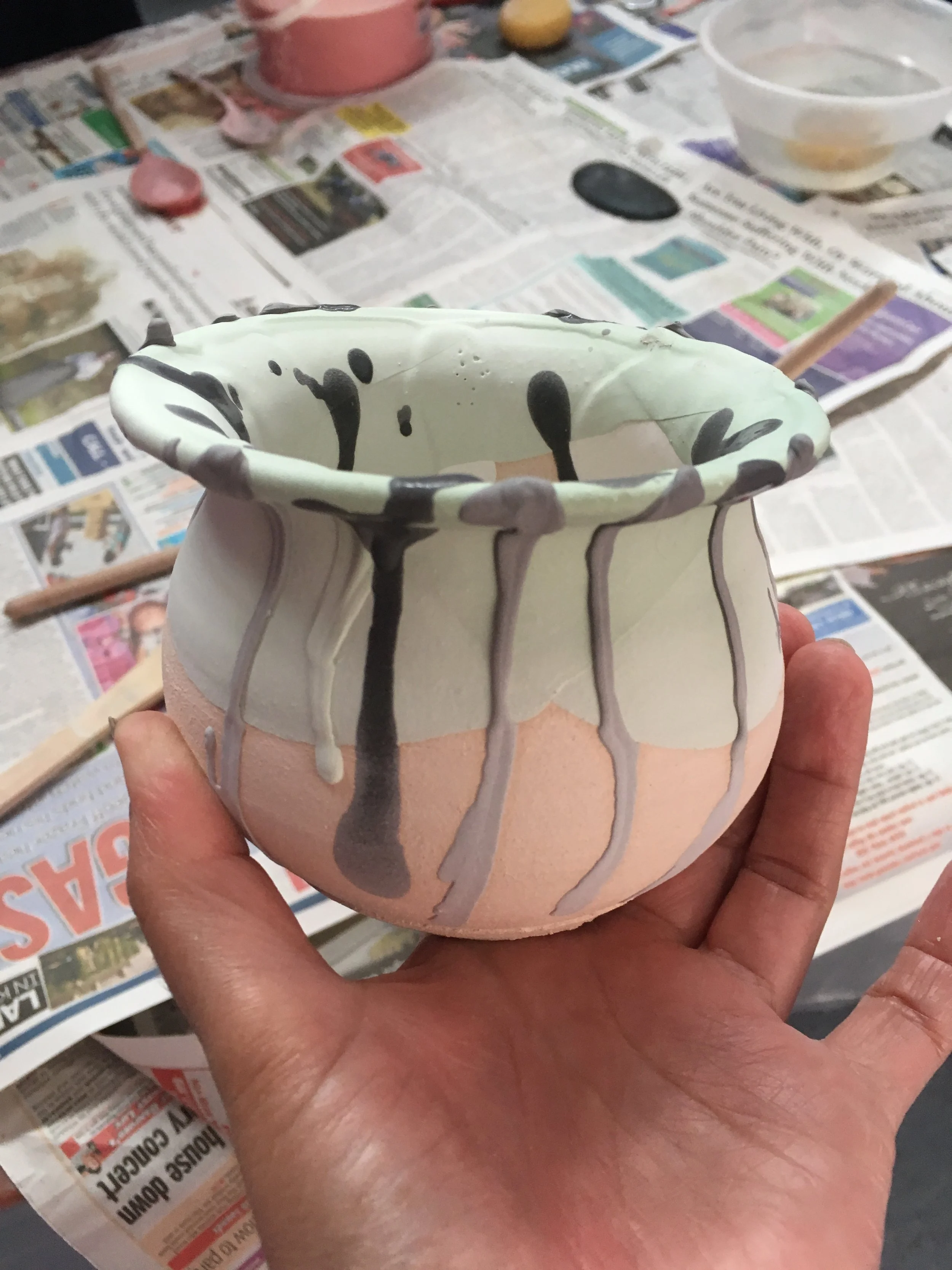 A hand holding a small ceramic cup with black and white abstract paint splatters, with some paint dripping down the sides, against a background of newspapers, paint brushes, and bowls of water for ceramic painting or glazing.