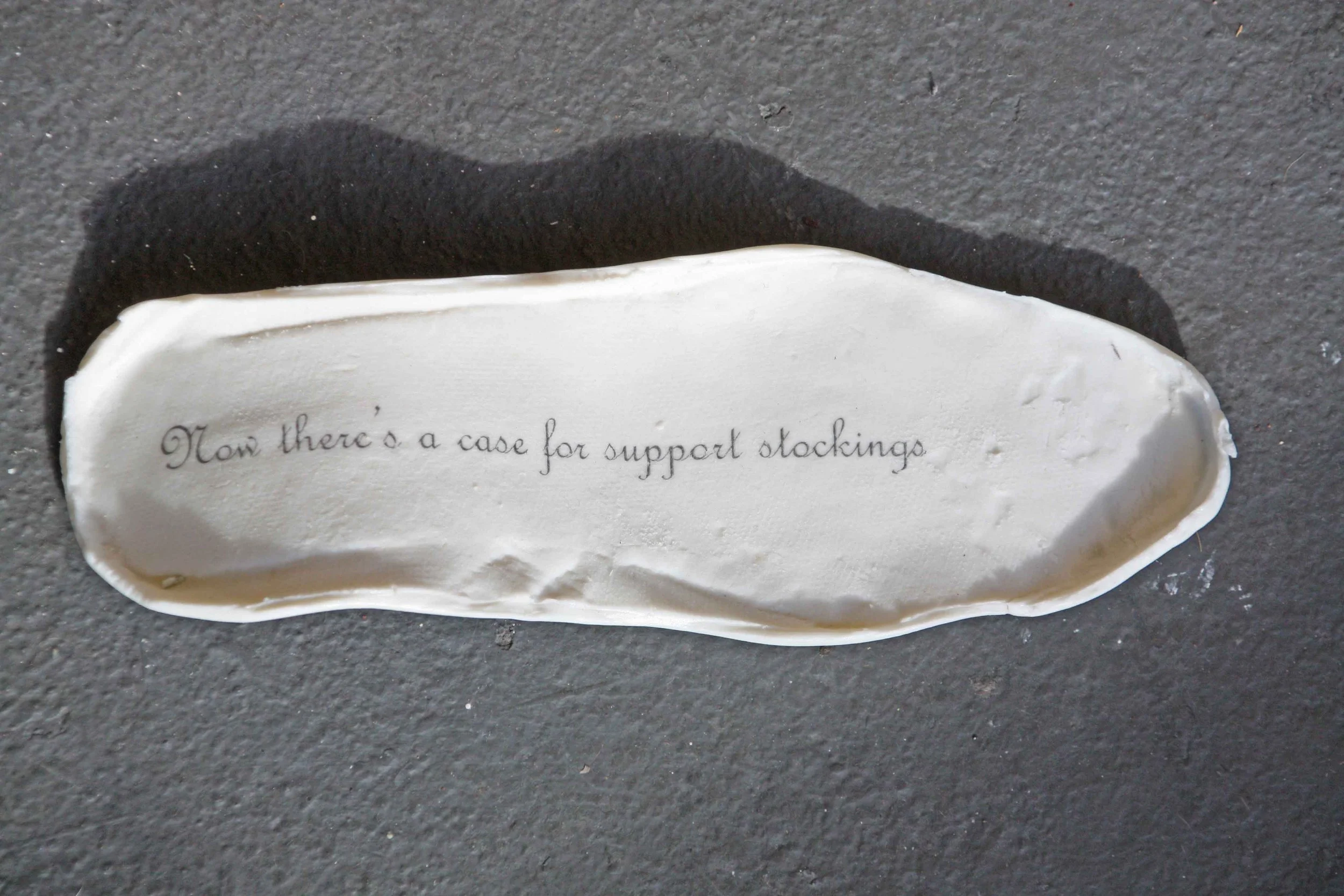 A white ceramic footprint with the phrase 'Now there's a case for support stockings' written on it, placed on a dark textured surface.