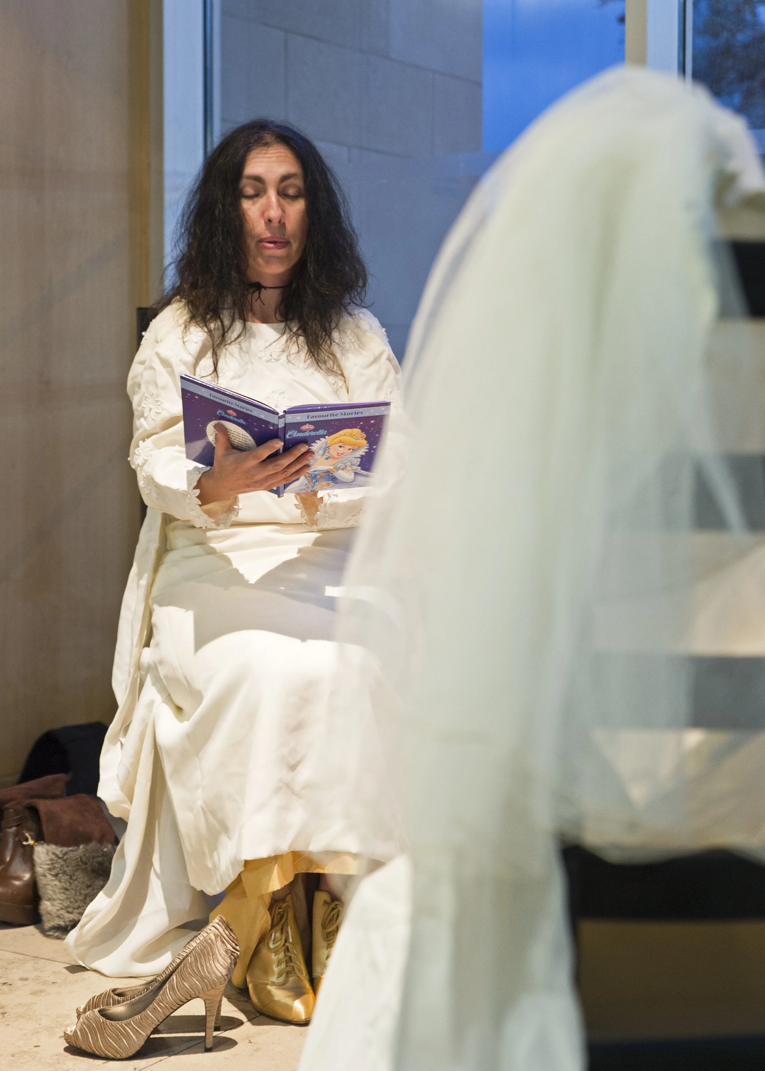 A woman with black, wavy hair reading a book, sitting indoors near a window with a stone wall behind her, surrounded by shoes including gold high heels, tan boots, and beige sneakers.