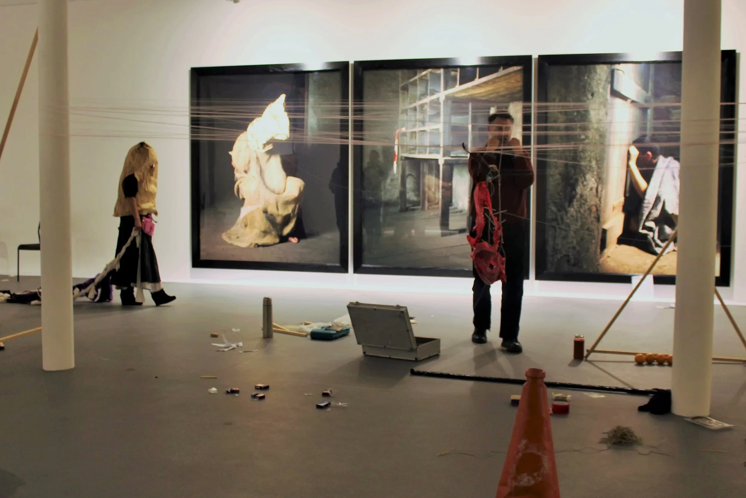 People working on an art installation or exhibit in a gallery or museum. Large framed photographs or artwork displayed on the wall with some objects and tools scattered on the floor.