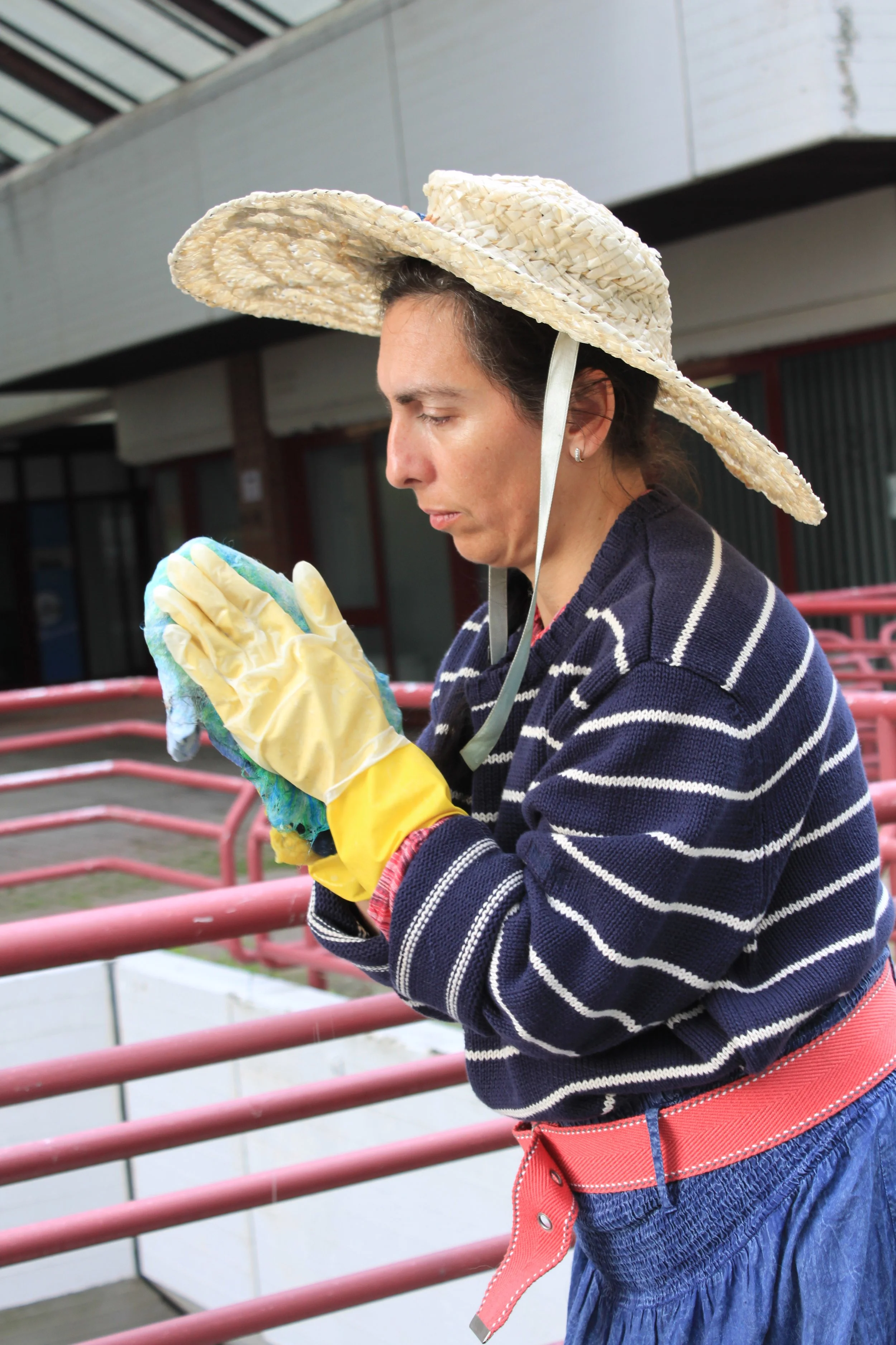 Woman wearing a large straw hat, yellow gloves, and a striped sweater, looking at her phone while standing near pink railings outdoors.