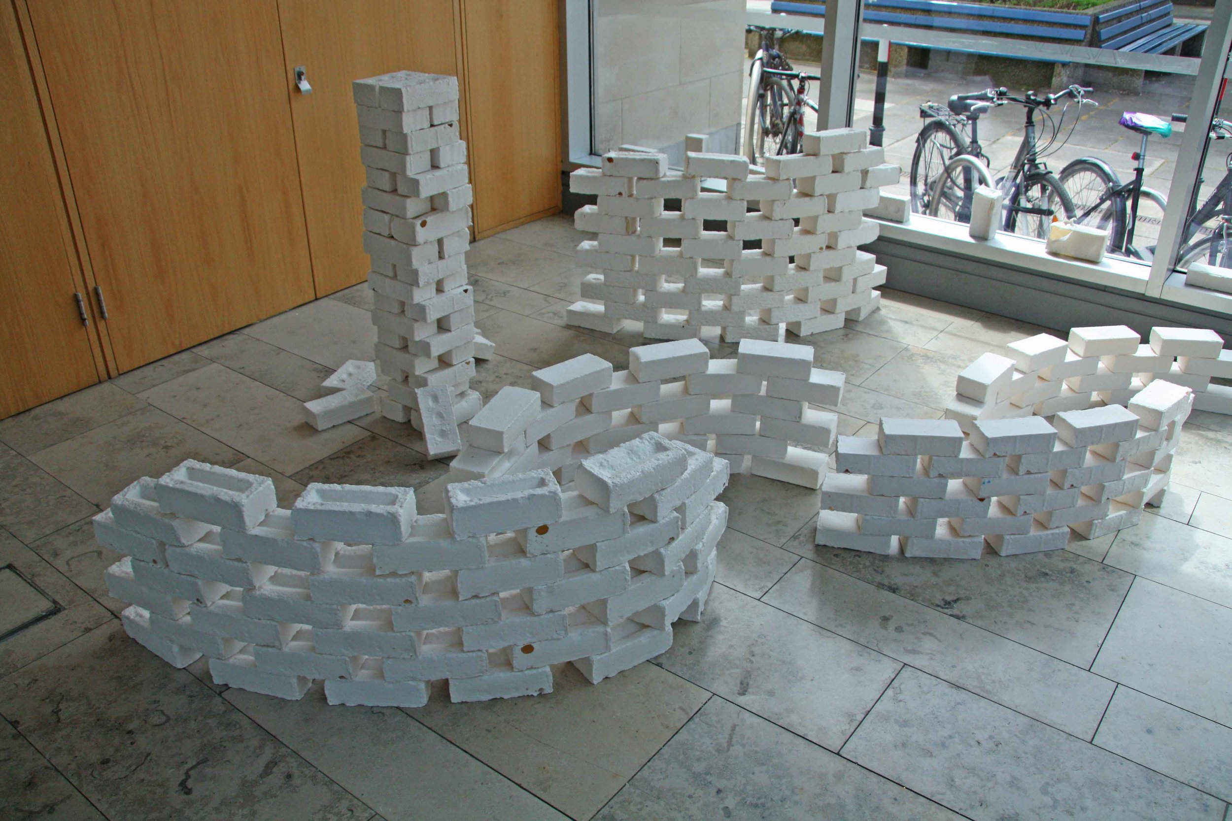 Indoor scene with white brick sculptures arranged on tiled floor, next to wooden doors and a large glass window with bicycles outside.
