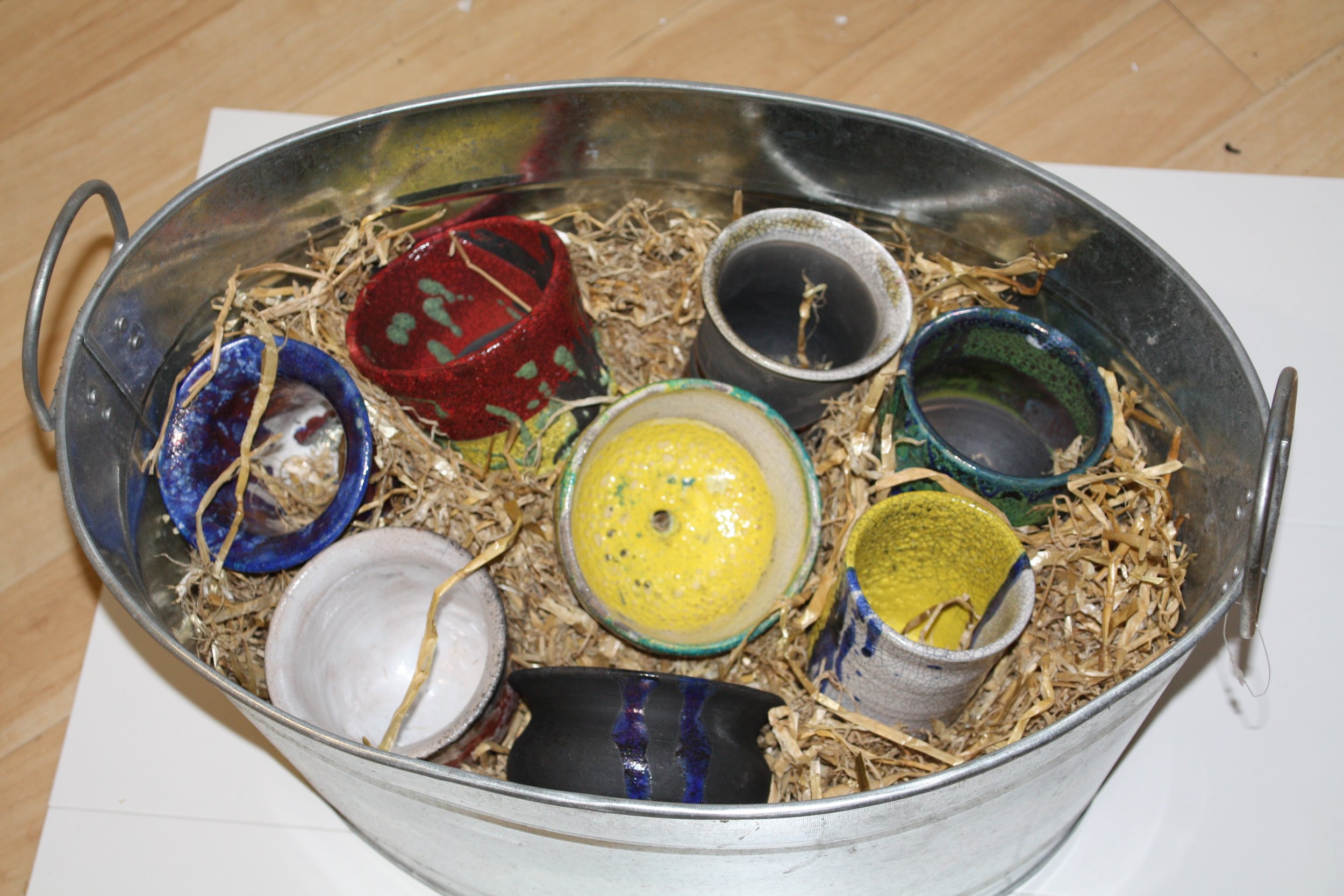 A metal bucket filled with shredded paper and multiple small, colorful ceramic cups, some of which are empty and some have yellow or dark-colored glaze, arranged on a flat surface.