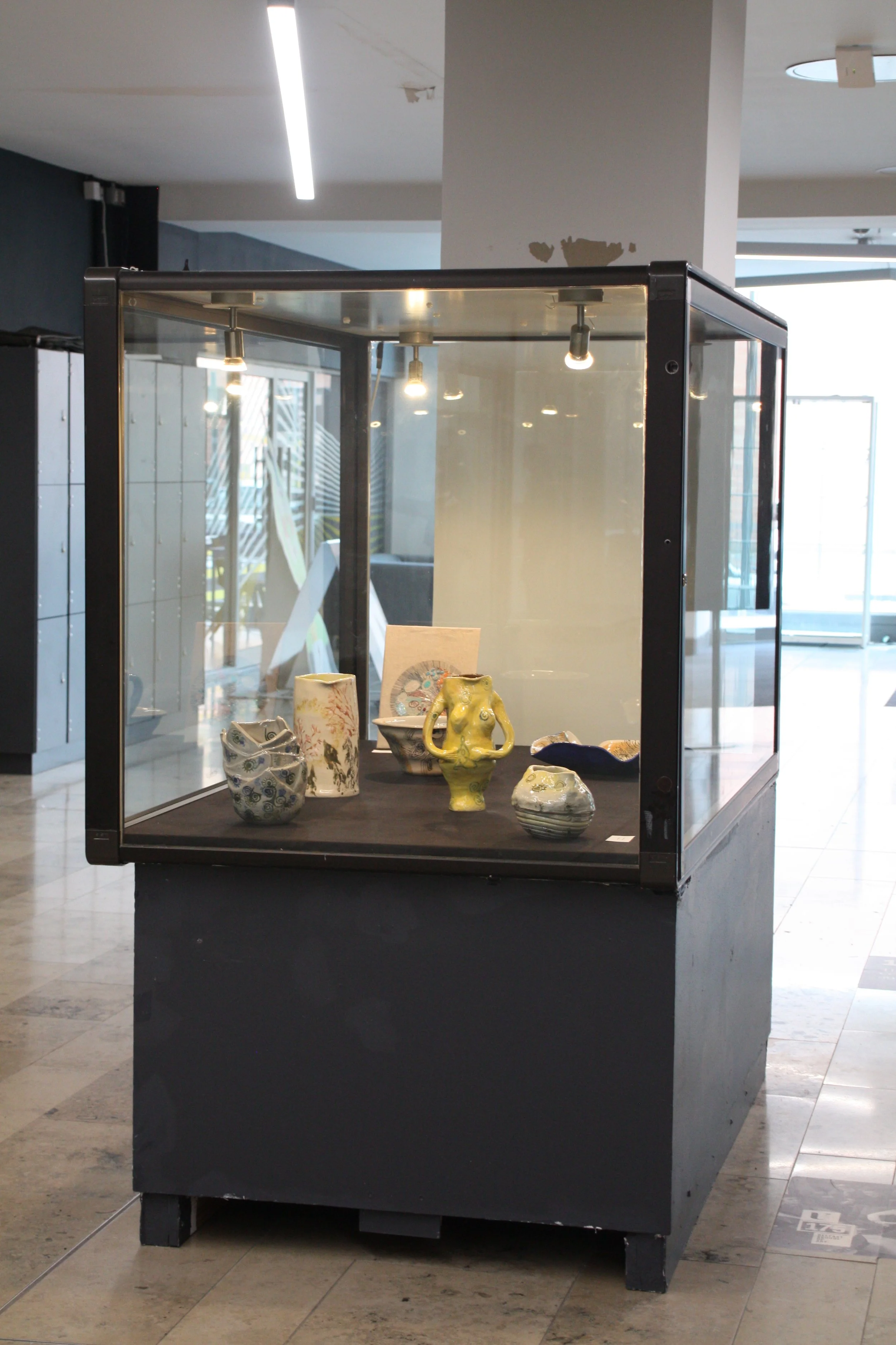 Glass display case showcasing various ceramic and pottery pieces, located inside a modern building with tiled flooring and overhead lighting.