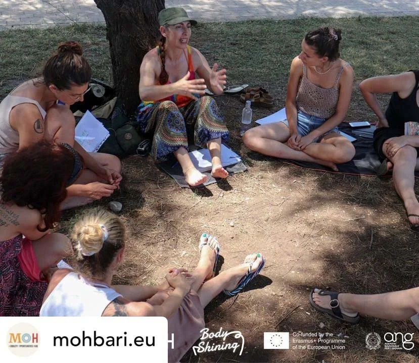 A group of women sitting in a circle on the ground outdoors, engaged in conversation. They are sitting under a tree, with some holding papers and others gesturing as they talk.