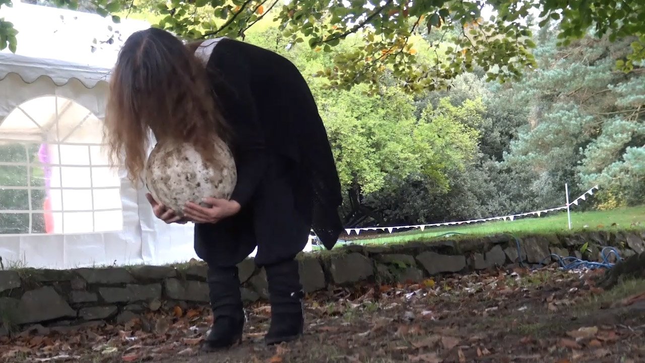 A person with long hair wearing dark clothing, holding a large speckled ceramic balls outdoors near a tent and trees.