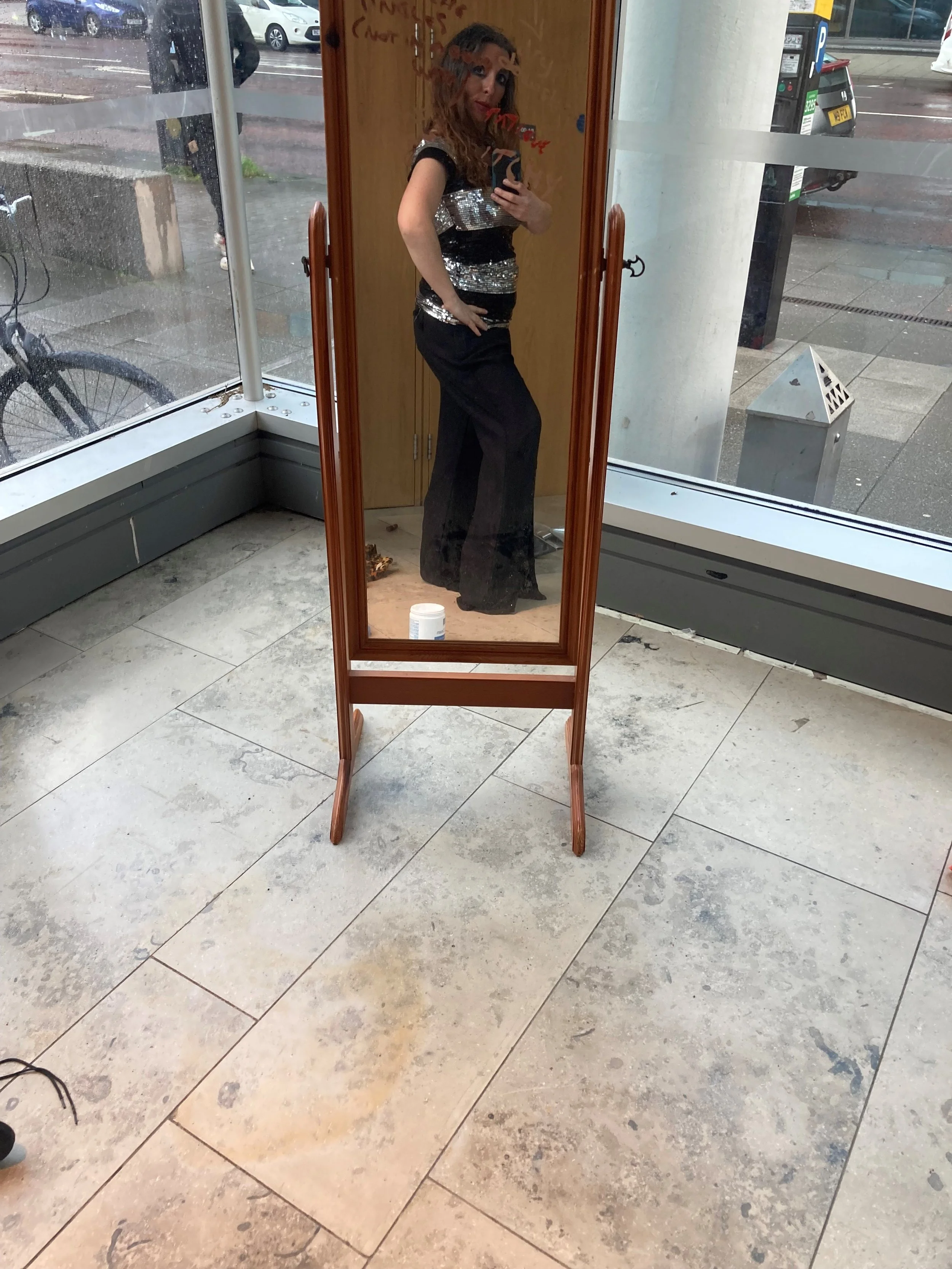 A woman with long curly hair wearing a shiny silver and black outfit is taking a mirror selfie inside a store. The store has large windows showing a rainy street outside. The reflection shows her posing with one hand on her hip and holding her phone 