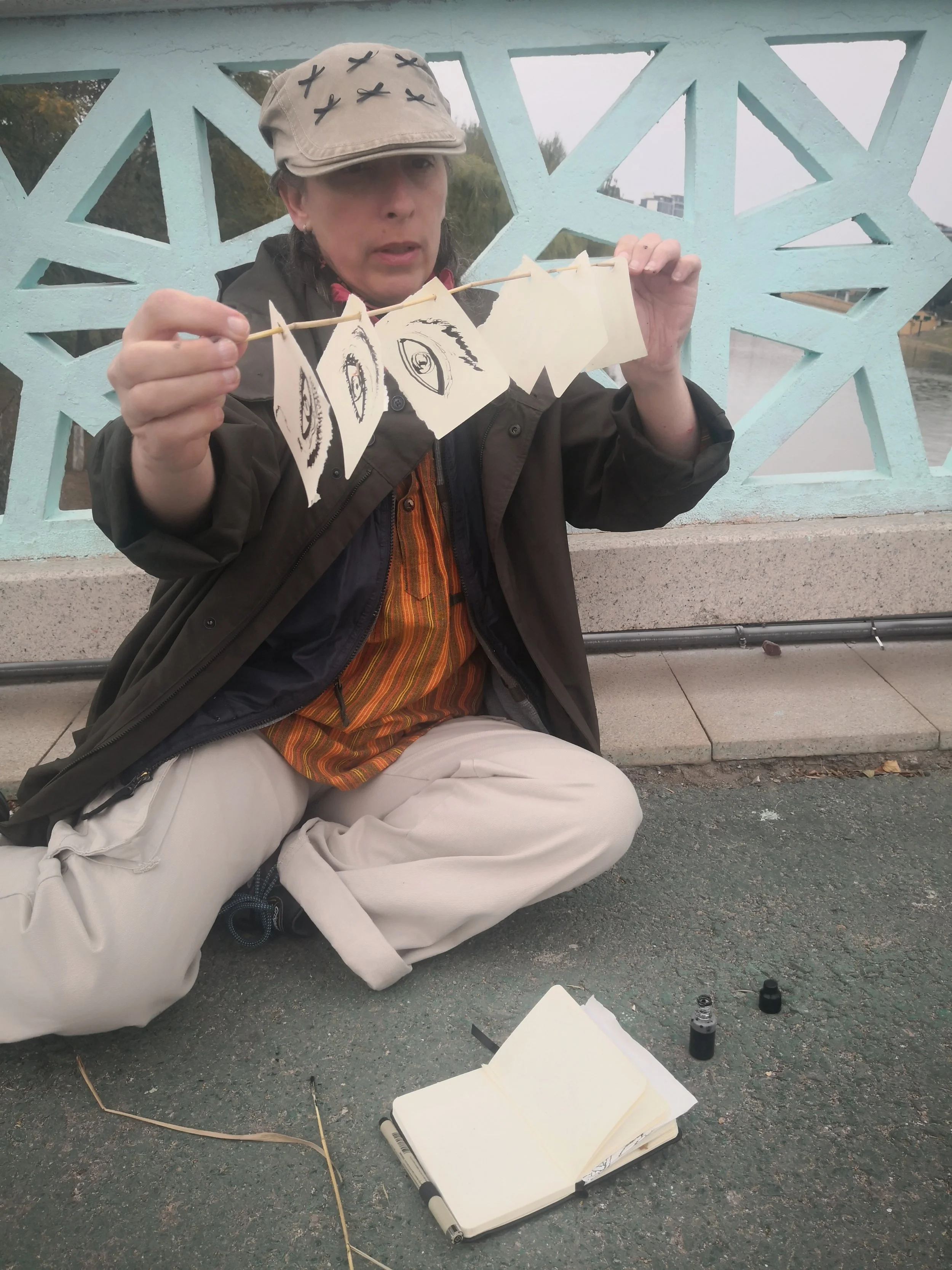 A woman sitting on the ground, holding a string with small art pieces attached, with an open sketchbook, bottles of ink, and markers nearby. She wears a beige cap with black designs and a dark jacket, with trees and a bridge in the background.