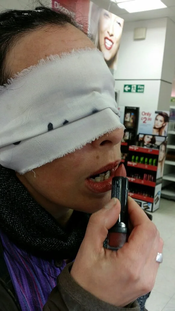 Person with a blindfold applying lipstick in a store aisle.