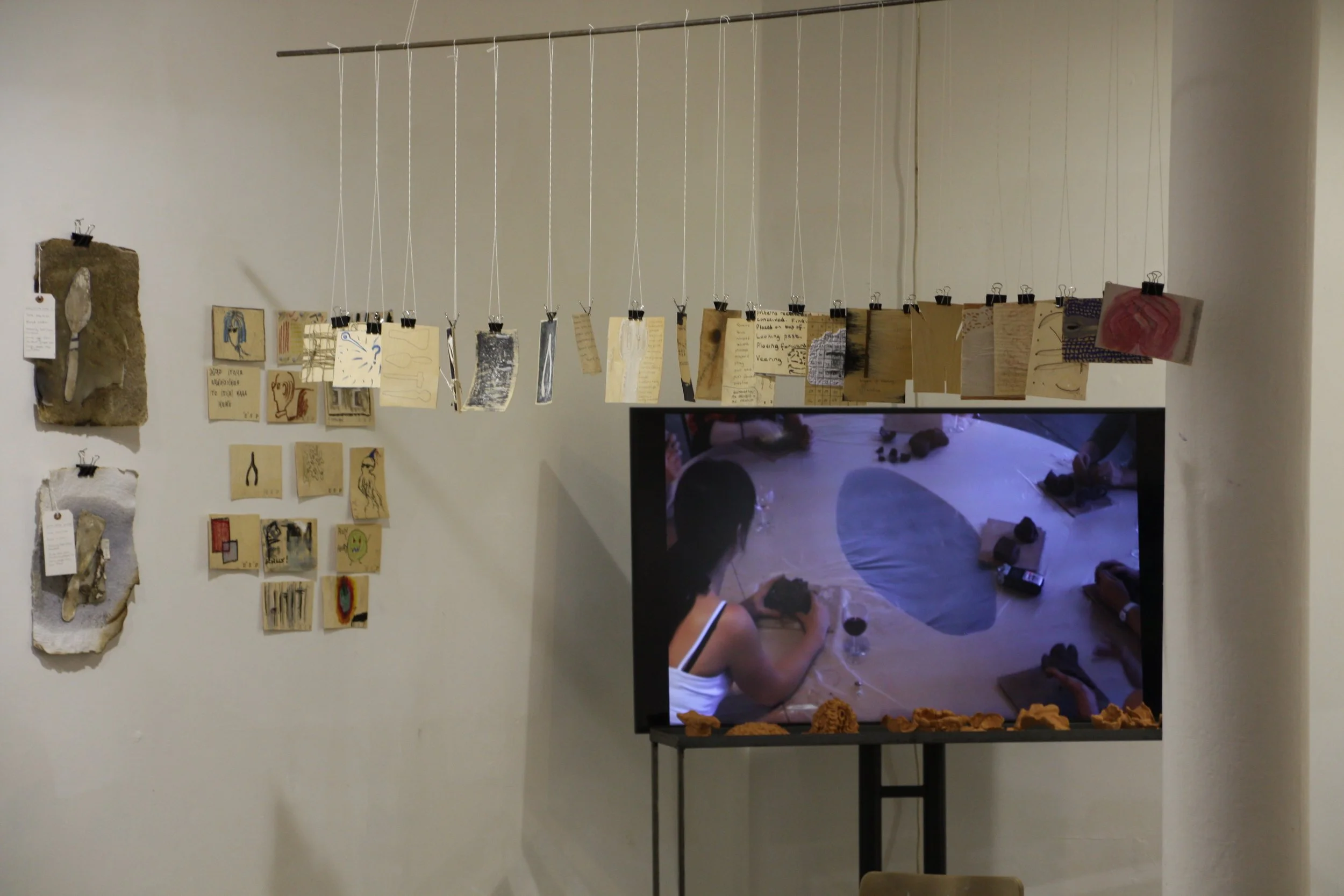 A wall decorated with various pieces of artwork, including sketches and textured pieces, and a string display with small hanging cards. A TV screen below shows people working on a craft project on the floor.