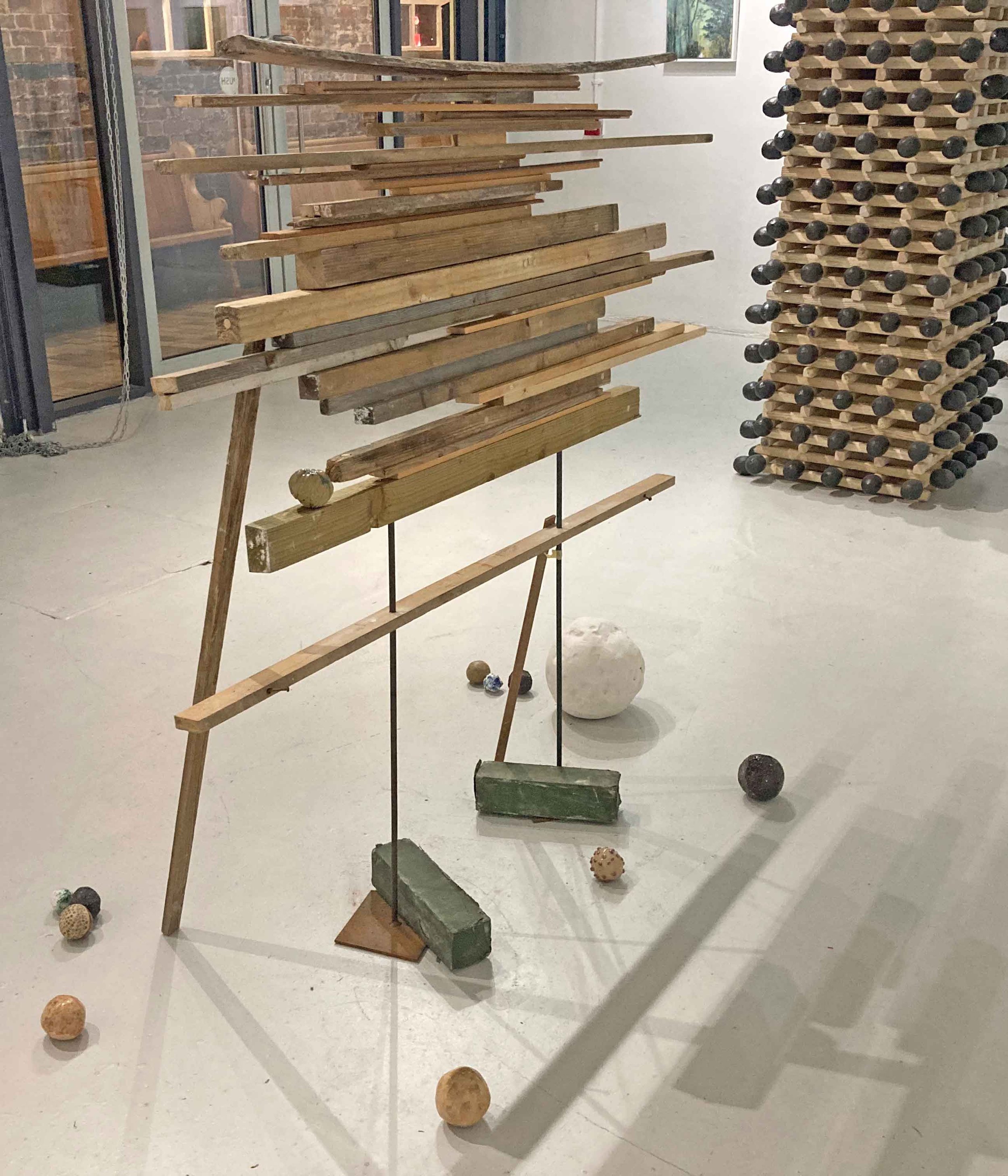 Art installation made of horizontal wooden planks and greenish bricks supported by vertical metal rods, displayed in an indoor gallery space.