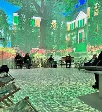 People sitting on benches in an interactive space that looks like an outdoor park area with trees and multi-story buildings in the background.