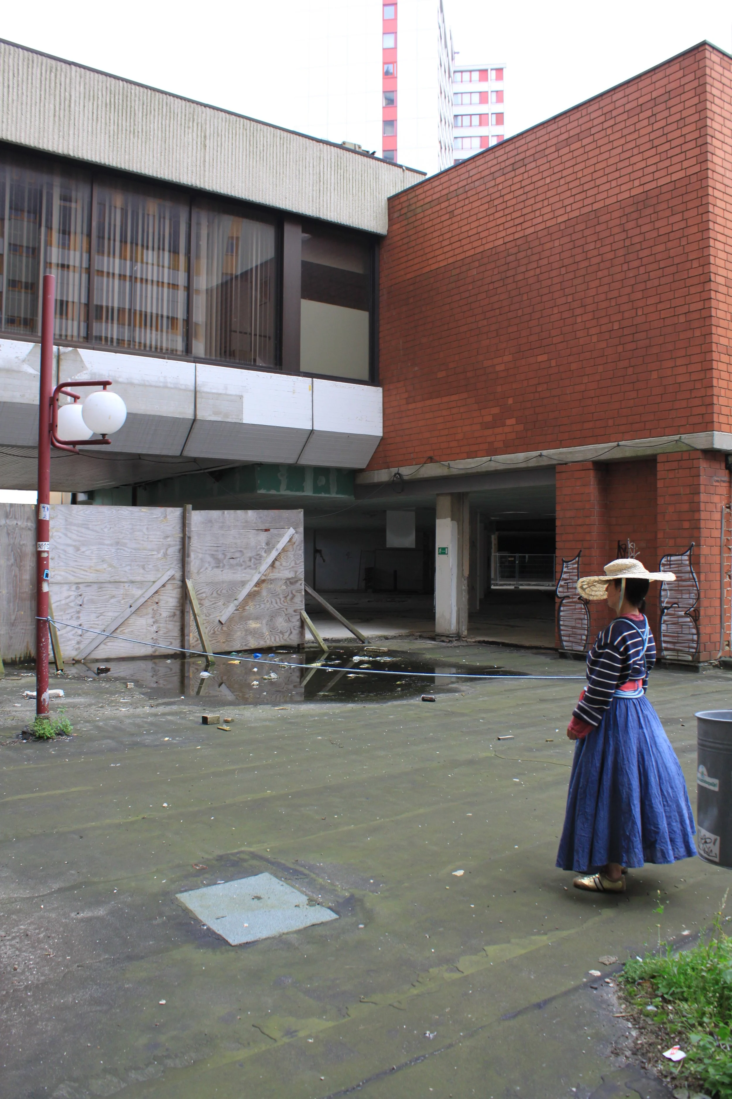 A woman wearing a wide-brimmed straw hat, striped top, and long blue skirt standing near a wet, trash-strewn pavement in an urban area with buildings, a construction barrier, and a light post nearby.