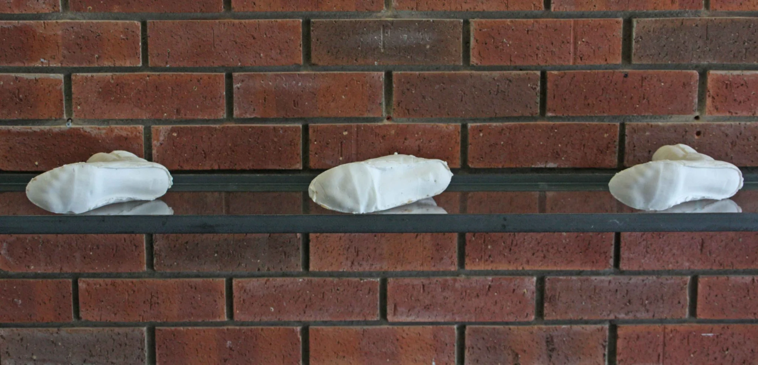 Three white inside shoe cast sculptures placed on a glass shelf with a red brick wall background.