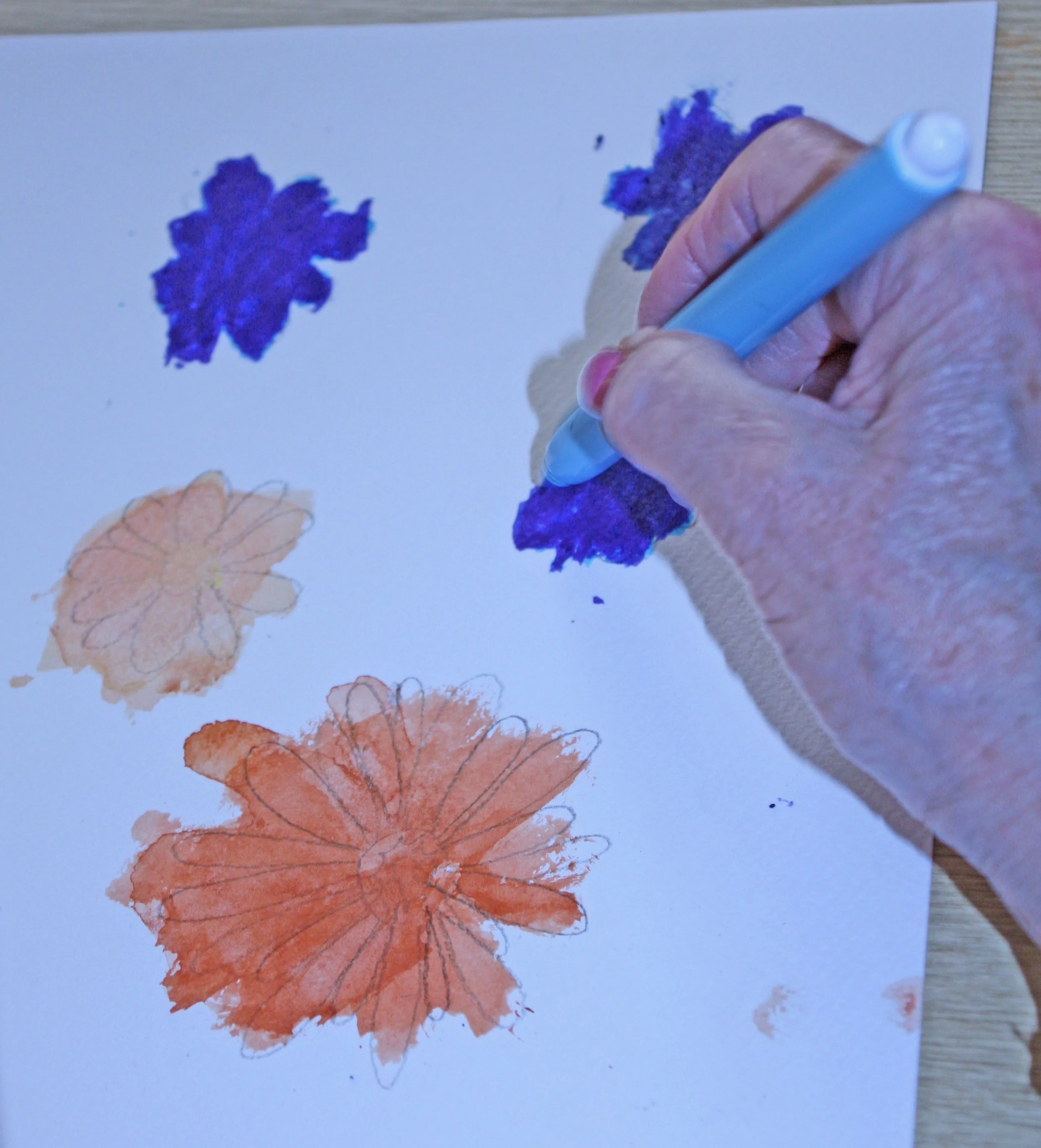 A person is using a blue pen to draw orange and purple leaf designs on white paper.