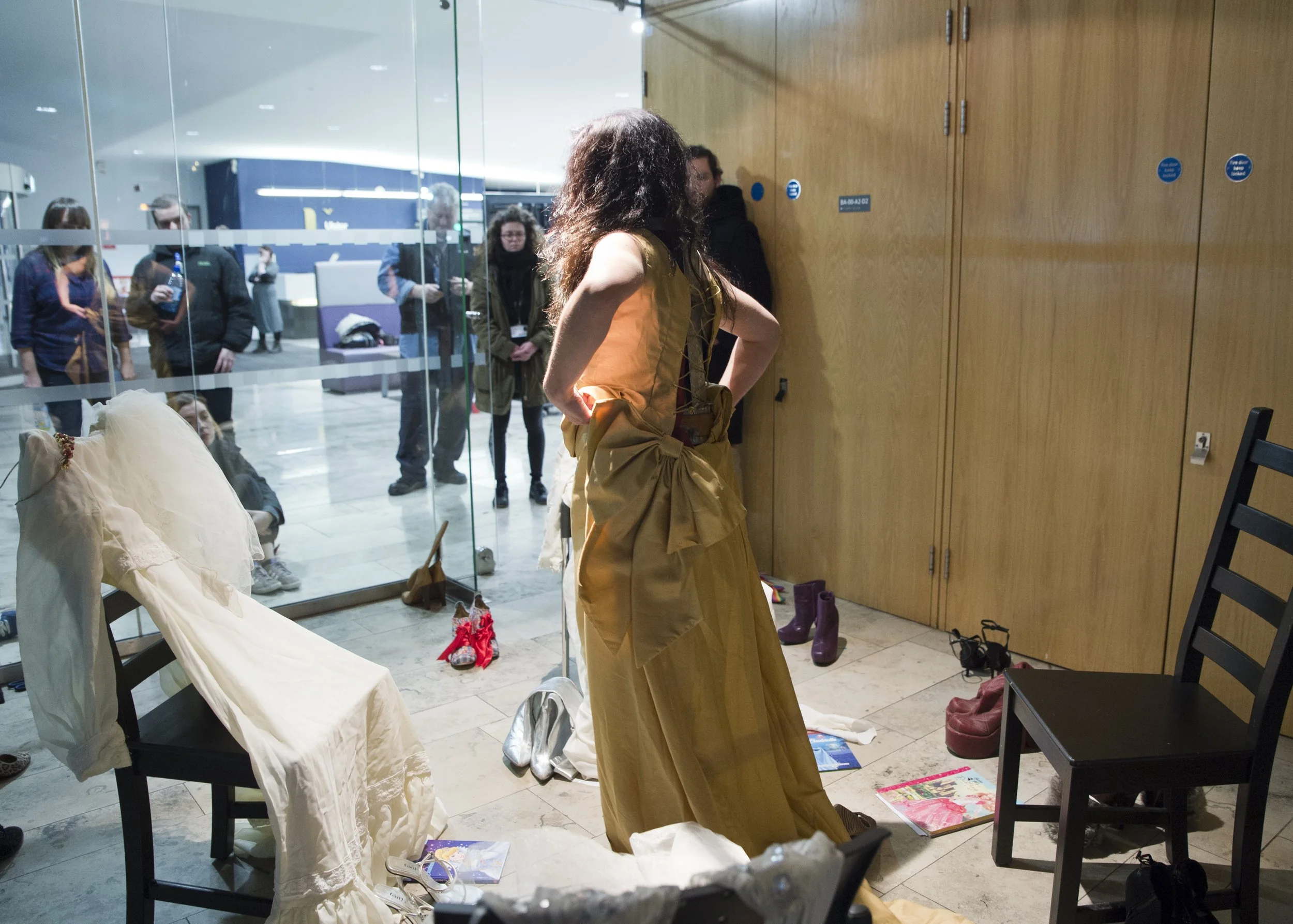 A woman with curly hair wearing a gold dress is standing with hand on her hip in front of a wooden wall. Behind her, there are other people in a waiting area with glass walls, and various shoes and magazines on the floor.
