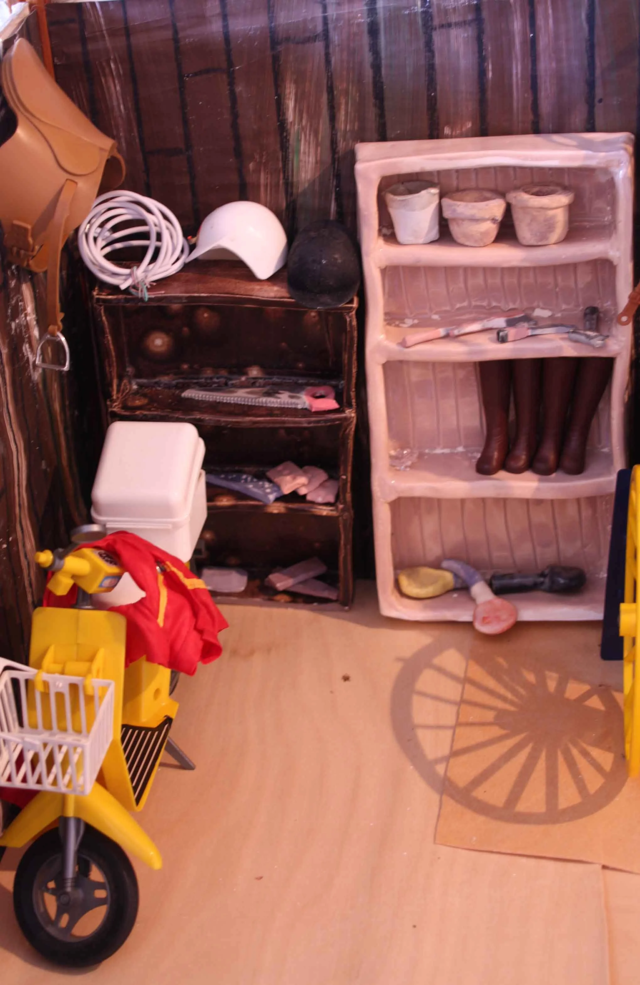 A shelf with three flower pots and a small objects, some brown boots. To the left, a white plastic container, a coiled white cord, a white helmet,. The floor is light-colored wood, and the wall behind is dark wood.