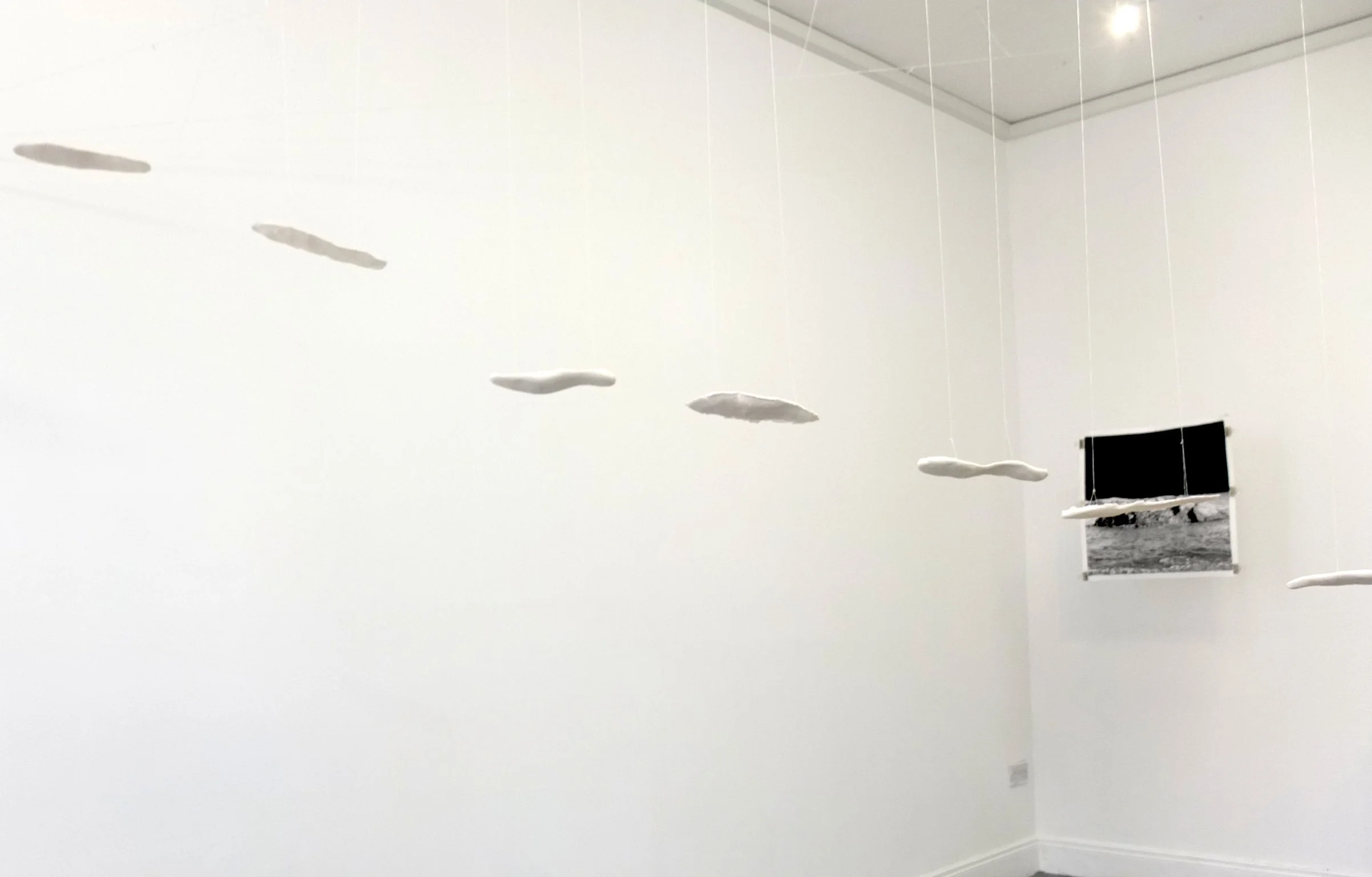 A white gallery wall with white footprints hanging  from thin wires and a black and white framed photograph on the floor.
