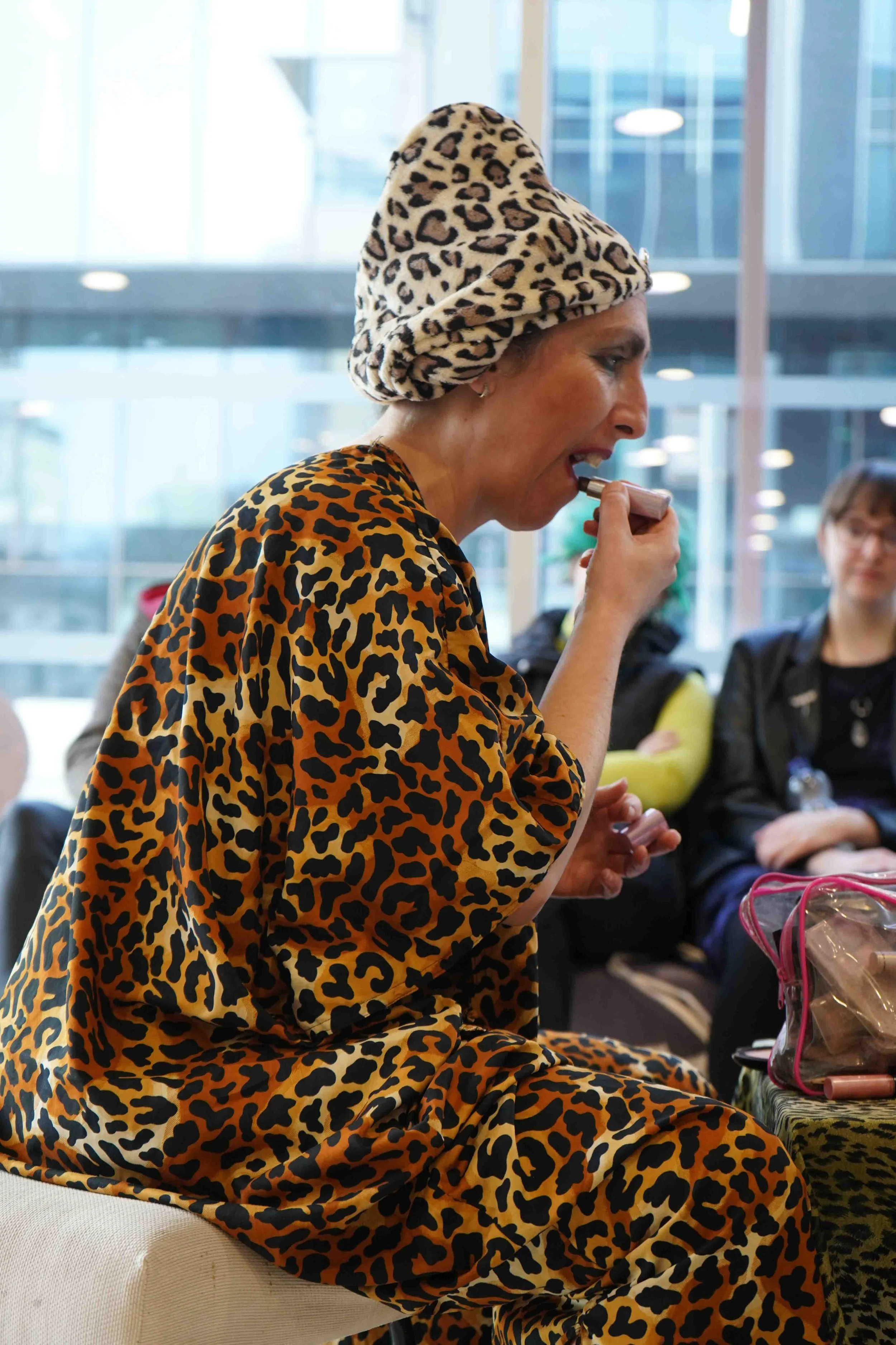 A woman is sitting in a, wearing a matching animal print outfit and a leopard print head covering. She is holding a lipstick or cosmetic item near her mouth, possibly applying makeup. There are other people in the background, some with bags or backpa