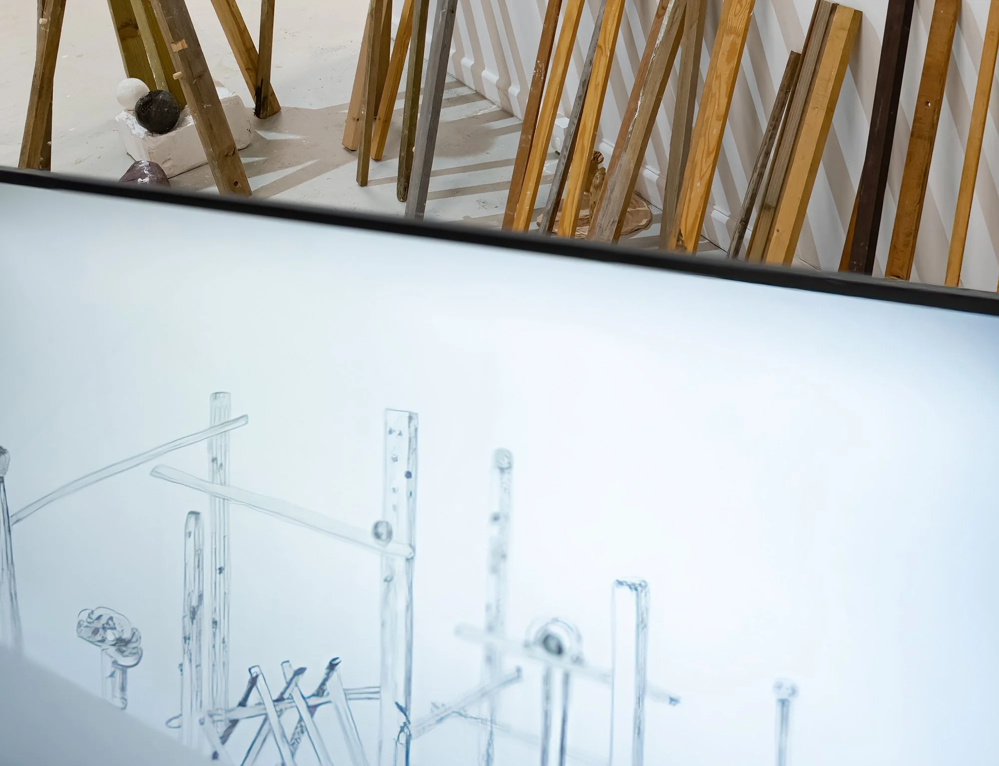 An art installation with vertical wooden slats arranged against a white wall, some supporting black and white spherical sculptures, and a transparent acrylic panel with a drawing of a chair and other objects.